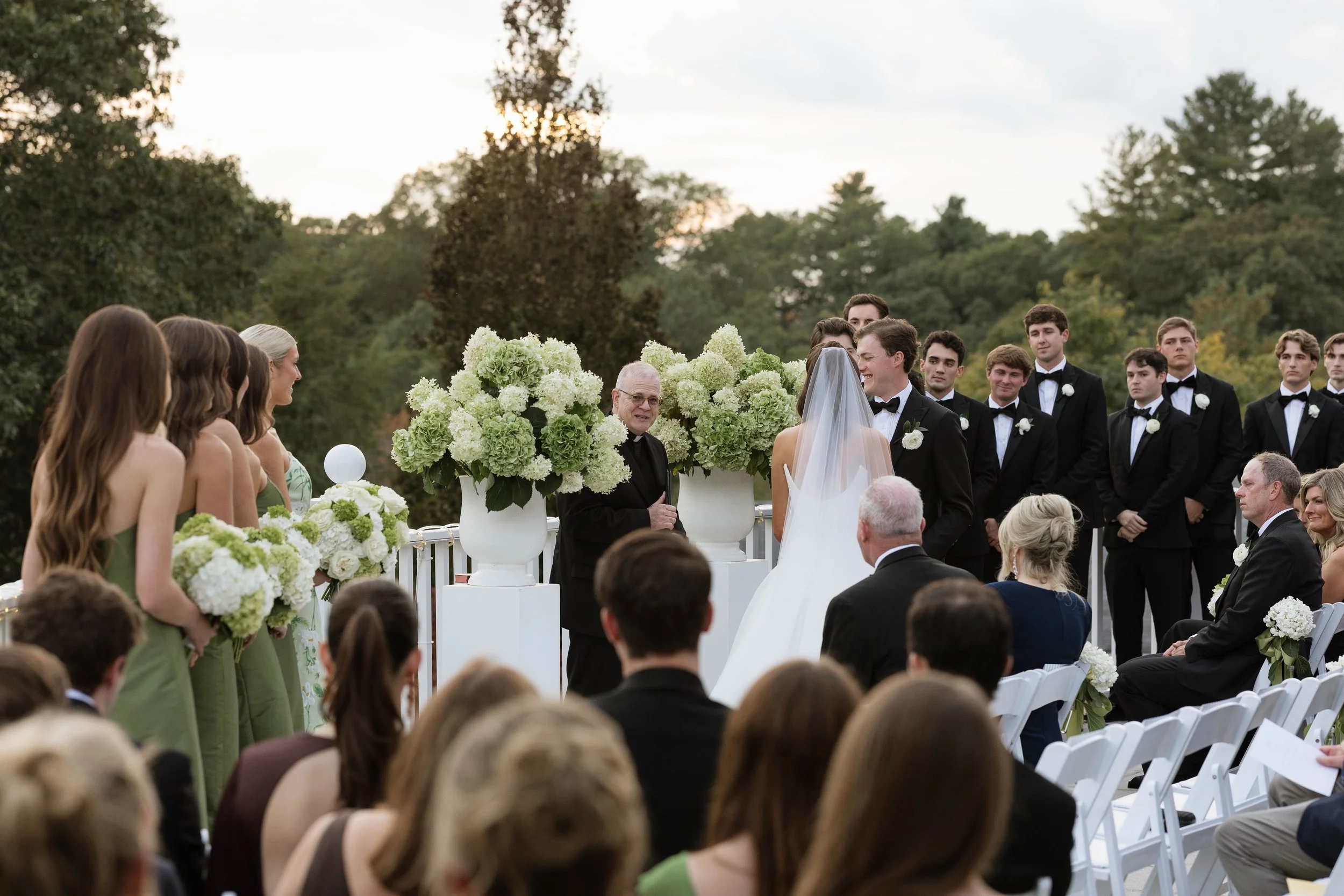 Outdoor wedding ceremony with bride and groom exchanging vows, surrounded by bridesmaids in green dresses and groomsmen in black tuxedos, set against trees and a cloudy sky.