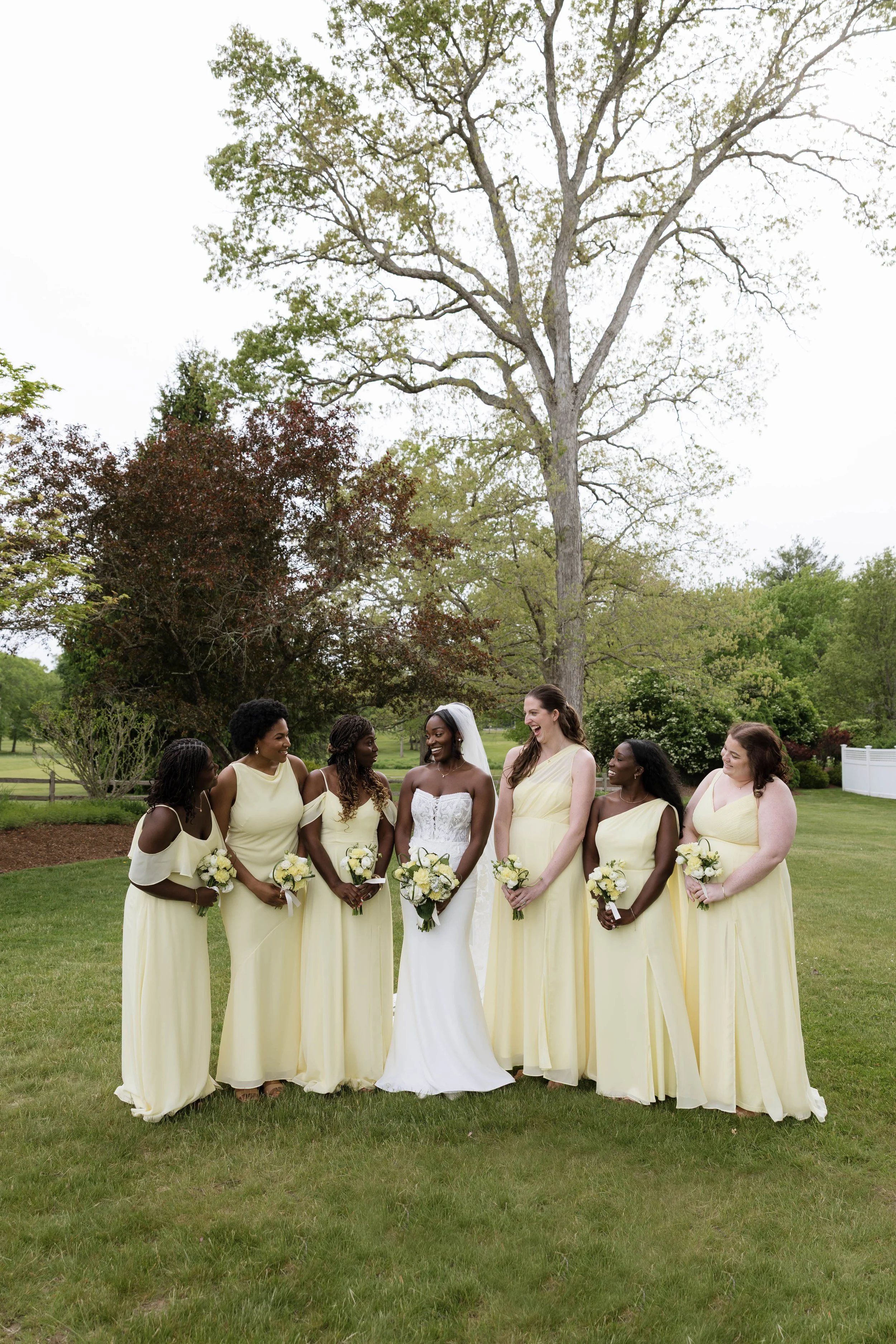 A group of women dressed in yellow bridesmaid dresses and a bride in a white gown, standing outdoors on a green lawn with trees and bushes in the background, some smiling and holding bouquets.