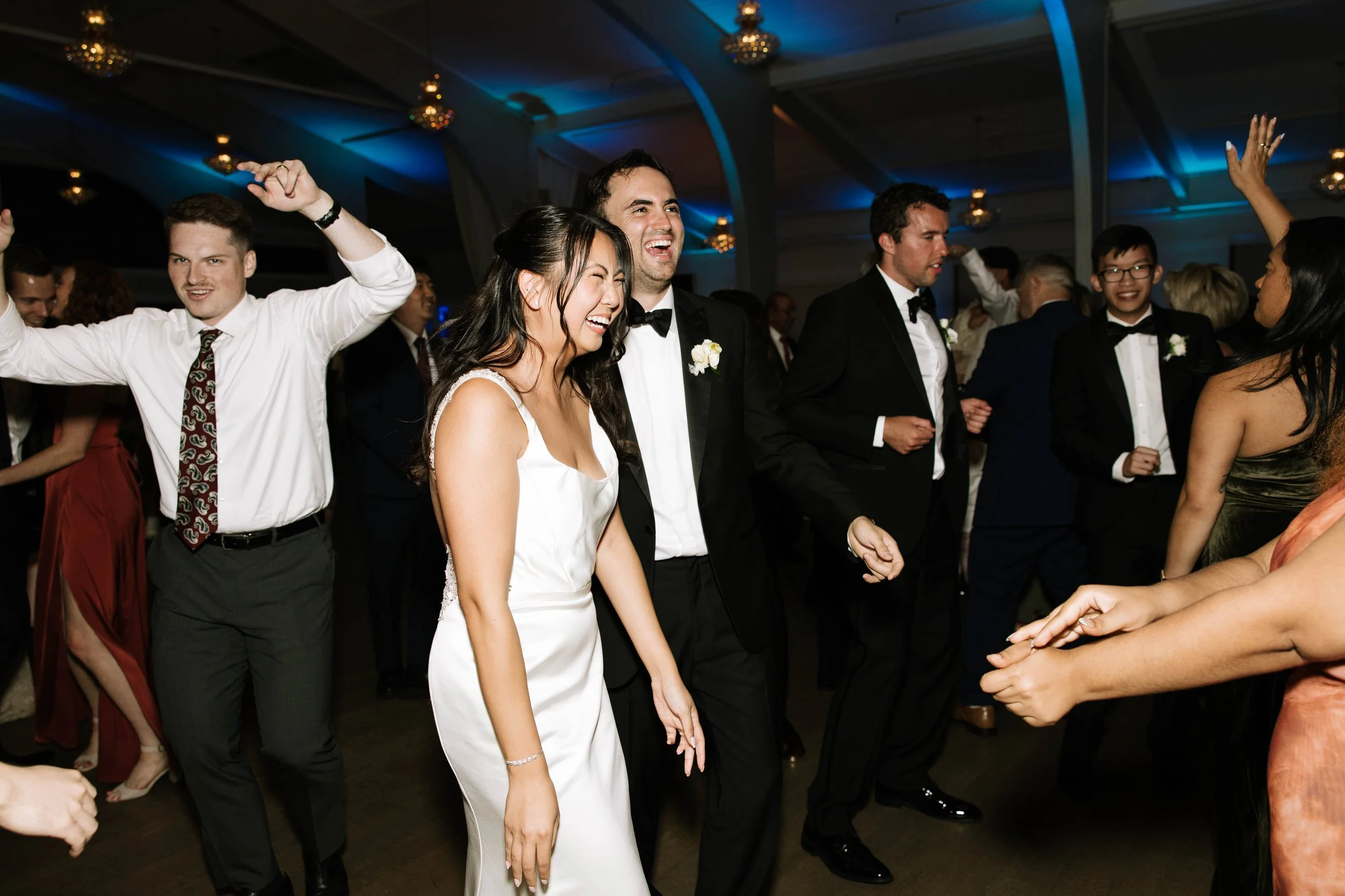 People dancing and celebrating at a wedding reception, with bride in a white dress and groom in a tuxedo, smiling and having fun.