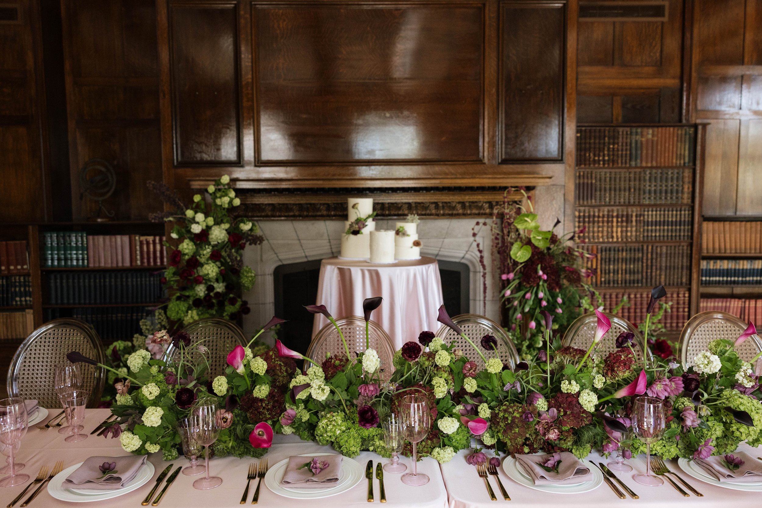 Elegant banquet table with pink tablecloth, floral centerpiece, and place settings, set in a room with wooden wall paneling, bookshelves, and floral arrangements.