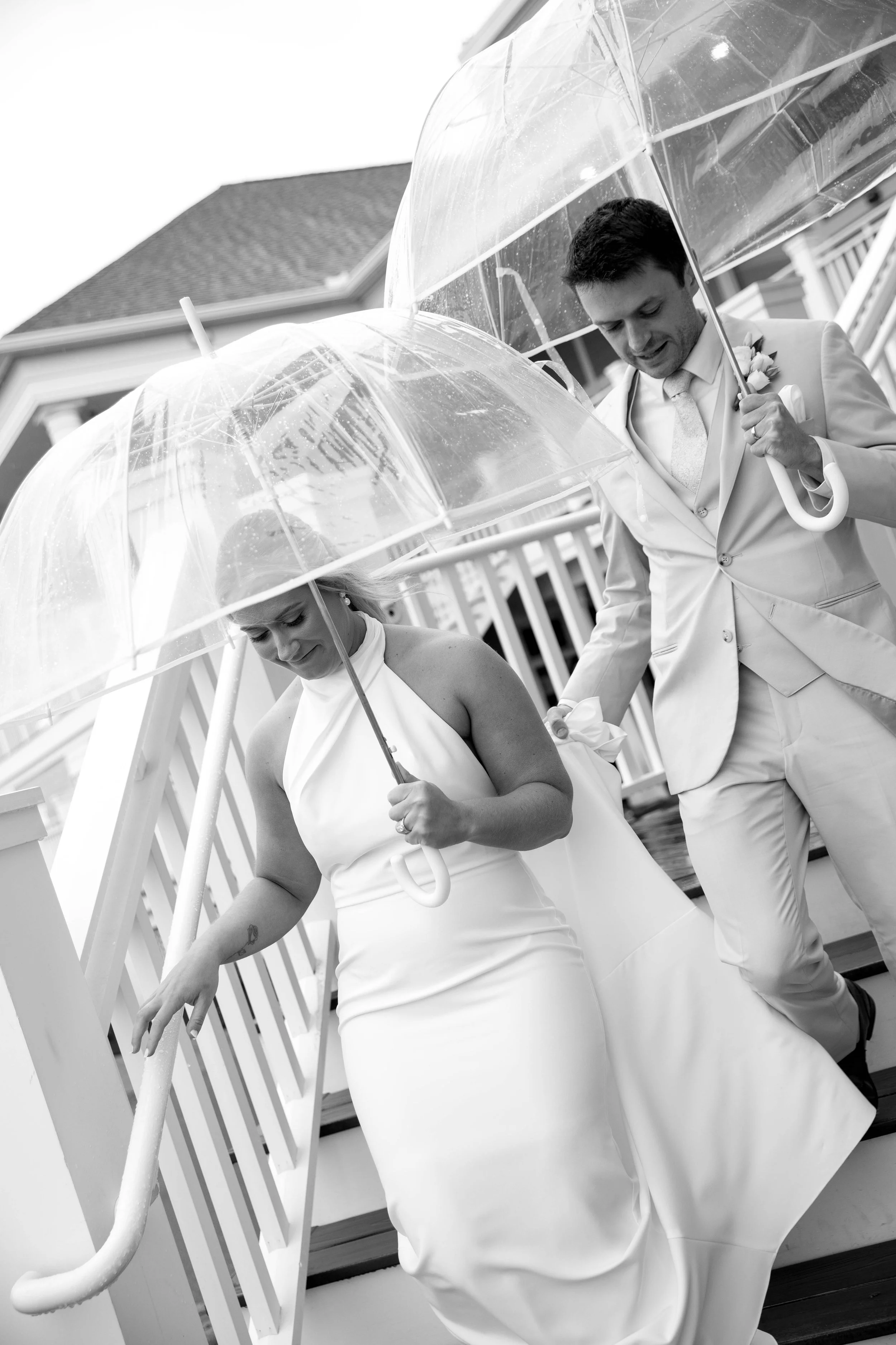 Black and white photo of a bride and groom walking down the stairs with umbrellas, dressed in wedding attire.