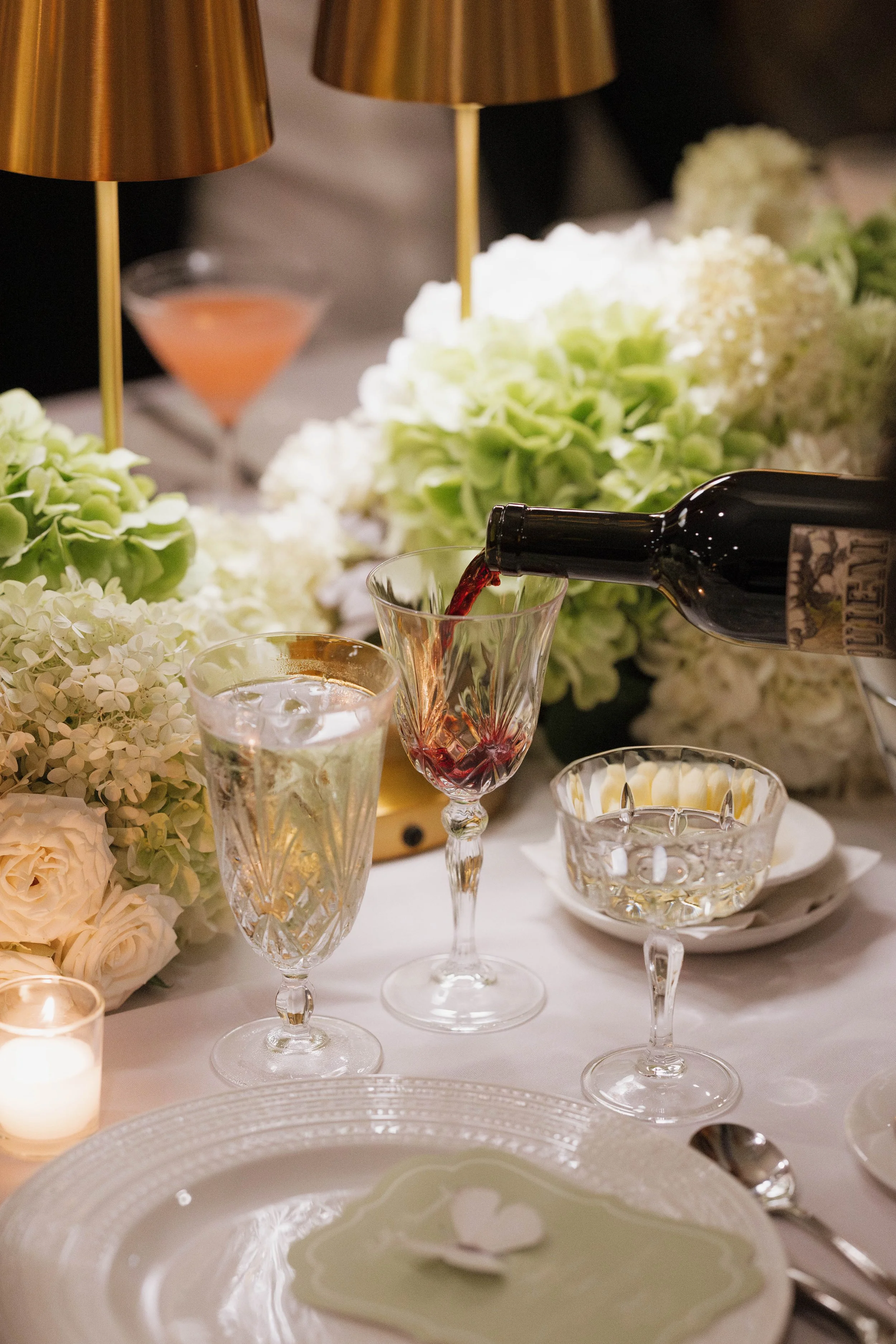 A table with glasses of red wine and water, surrounded by white and green floral arrangements, and a candle.