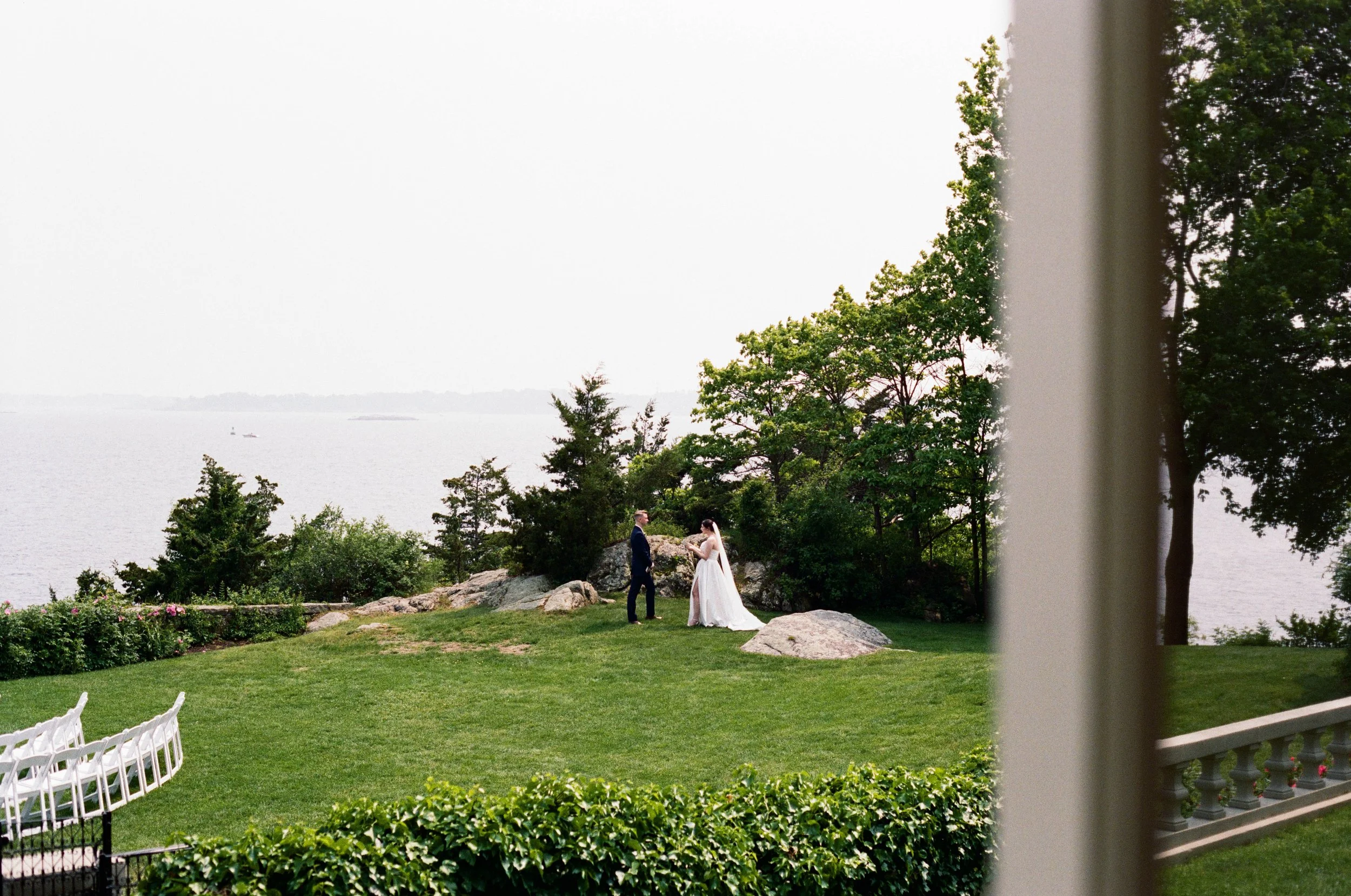 A bride and groom standing outdoors on a grassy area with rocks and trees near a large body of water, viewed from a window with a partially visible white column on the right side.