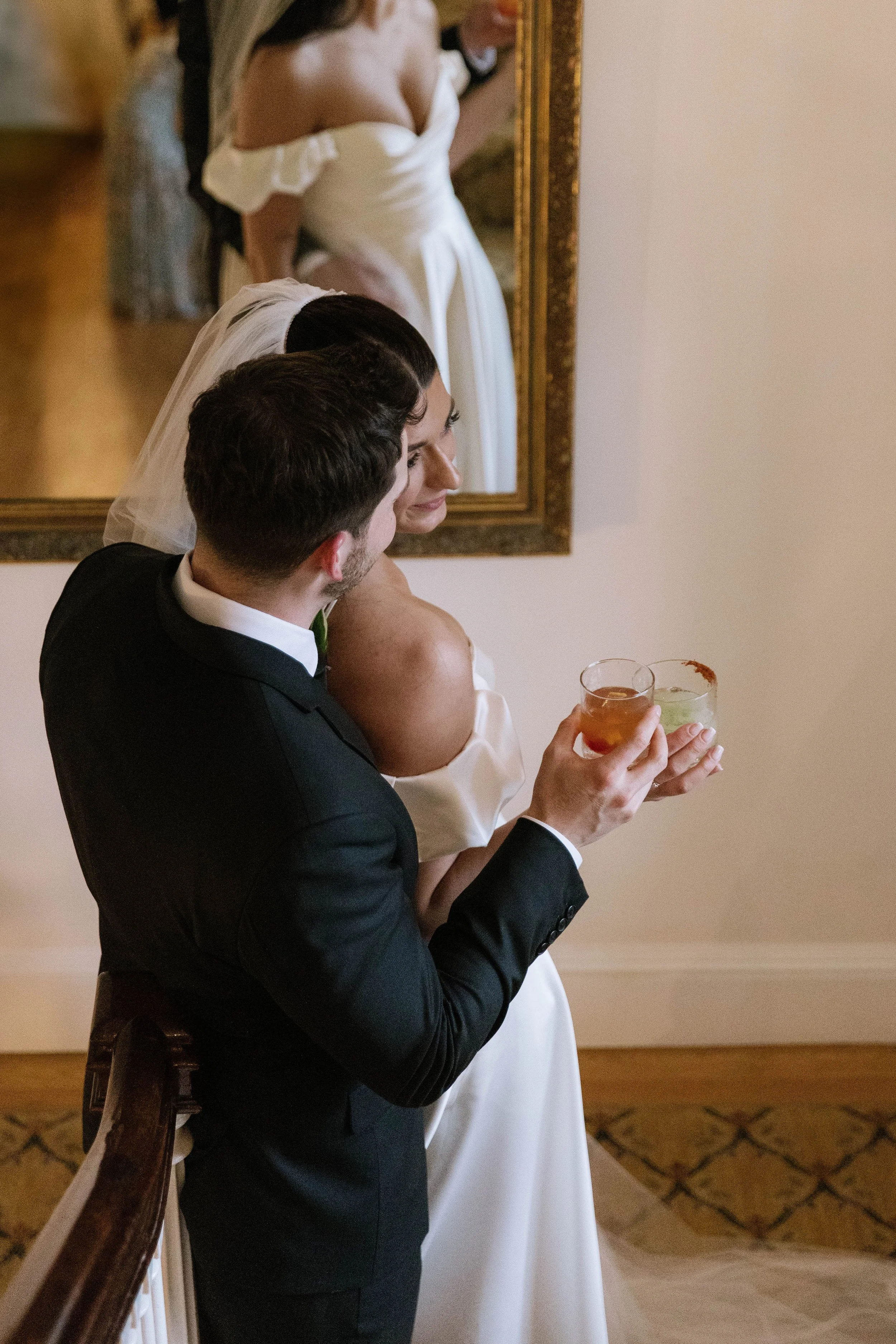 A bride and groom sharing a toast at a wedding, with a woman in a white dress and veil visible in the mirror behind them.