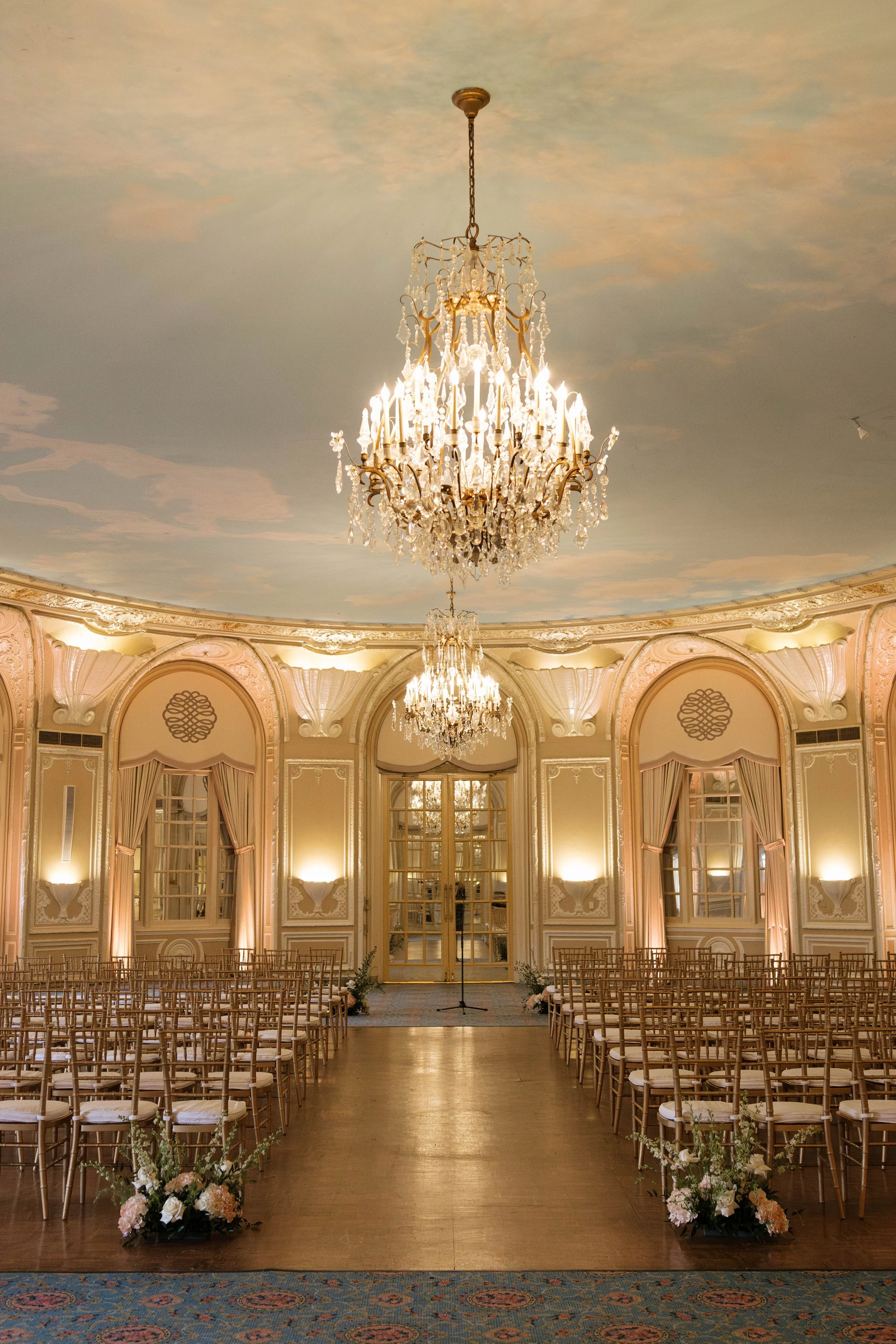 Elegant ballroom decorated for a wedding ceremony with a large chandelier, floral arrangements on the aisle, and rows of chairs facing a microphone stand at the front.
