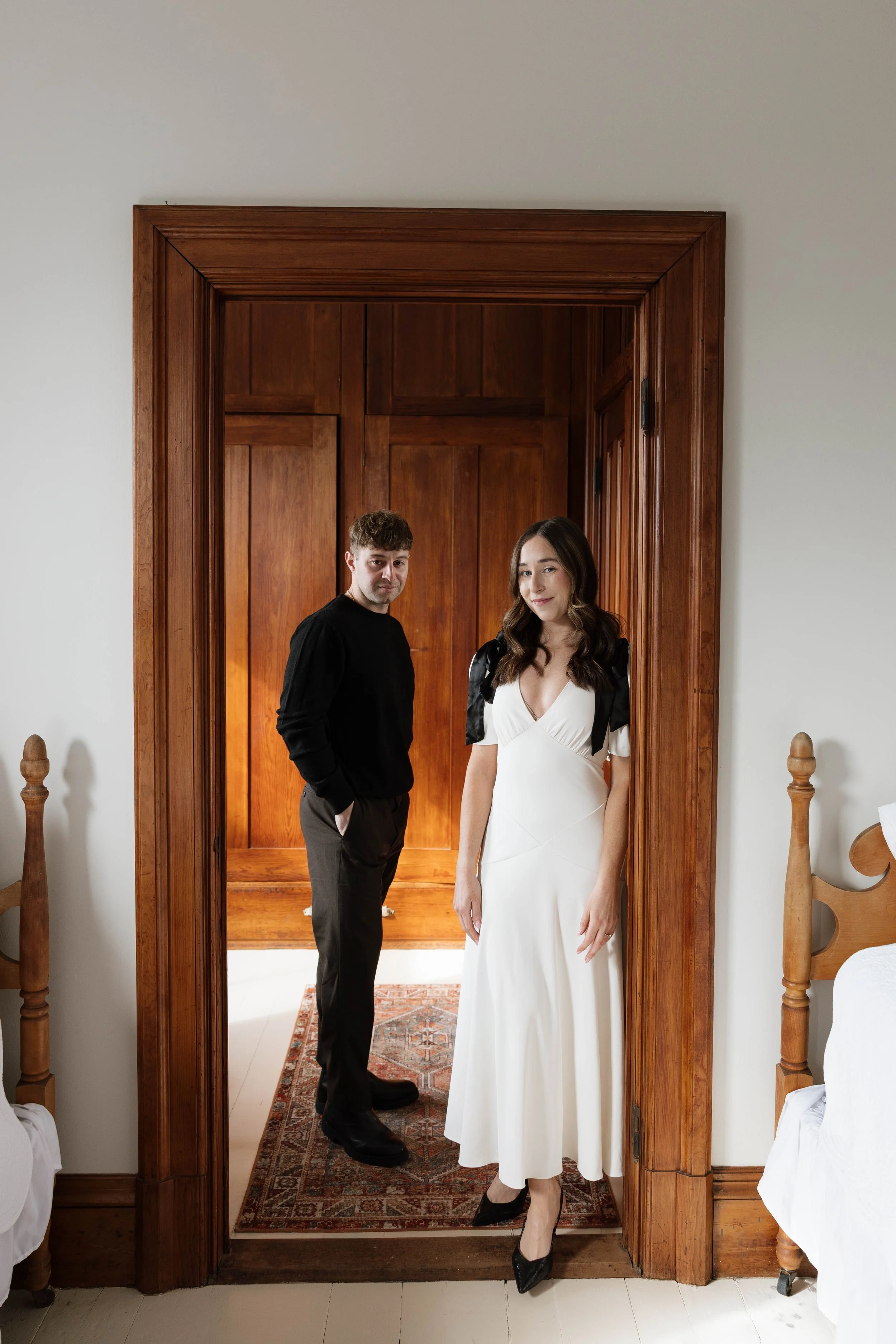 A man and woman standing in a doorway inside a room with wooden trims. The woman is wearing a white dress and black heels, while the man is in a black sweater and dark pants.