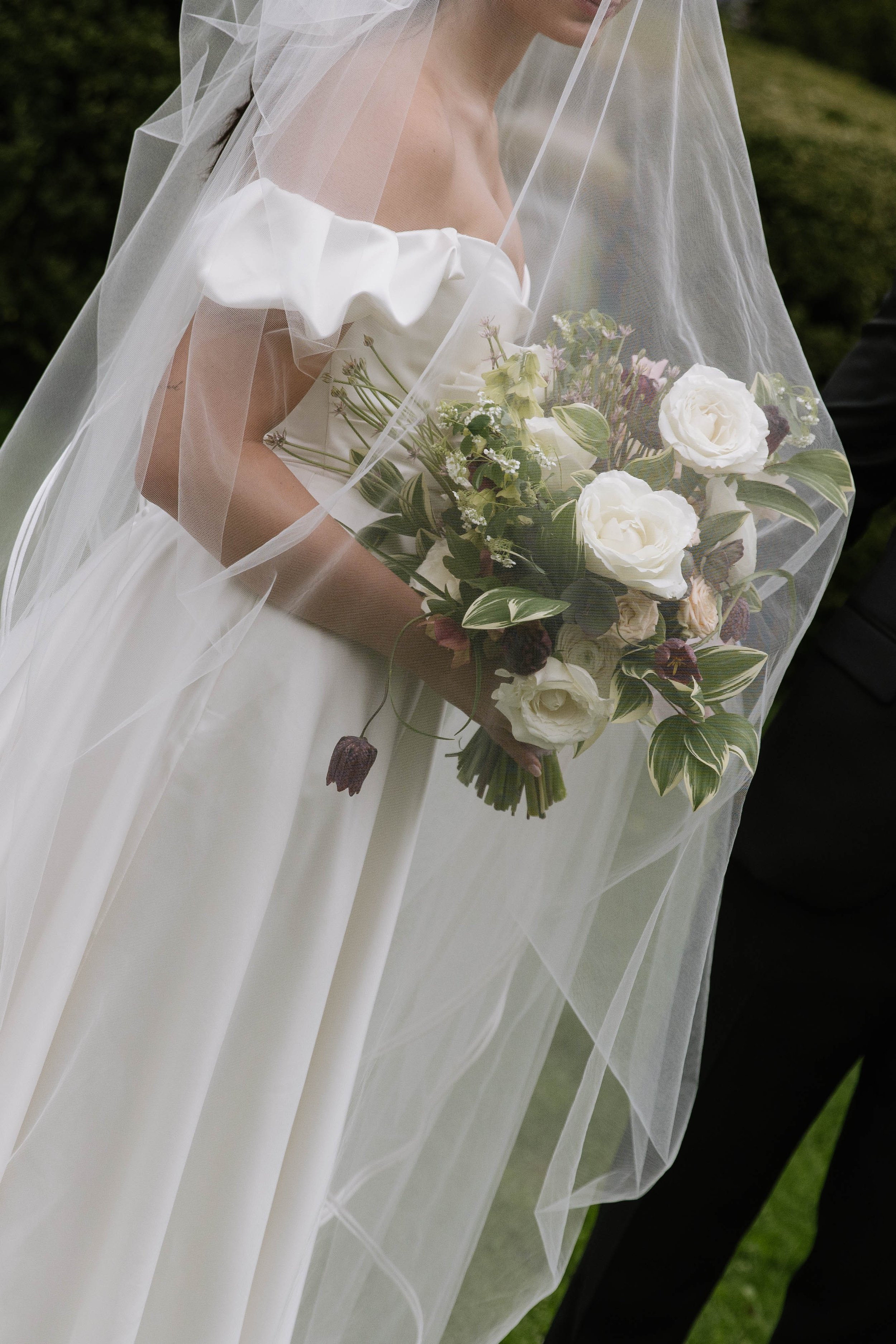 Close-up of a bride holding a bouquet of white and pink flowers on her wedding day, wearing a white off-shoulder dress and veil, outdoors with green grass in the background.
