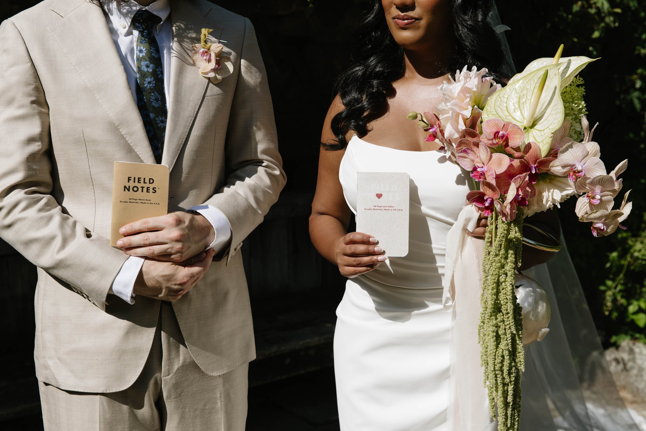 A bride and groom at their wedding ceremony, holding small cards and standing outdoors. The bride is holding a bouquet of pink and white flowers, and the groom is dressed in a light-colored suit.