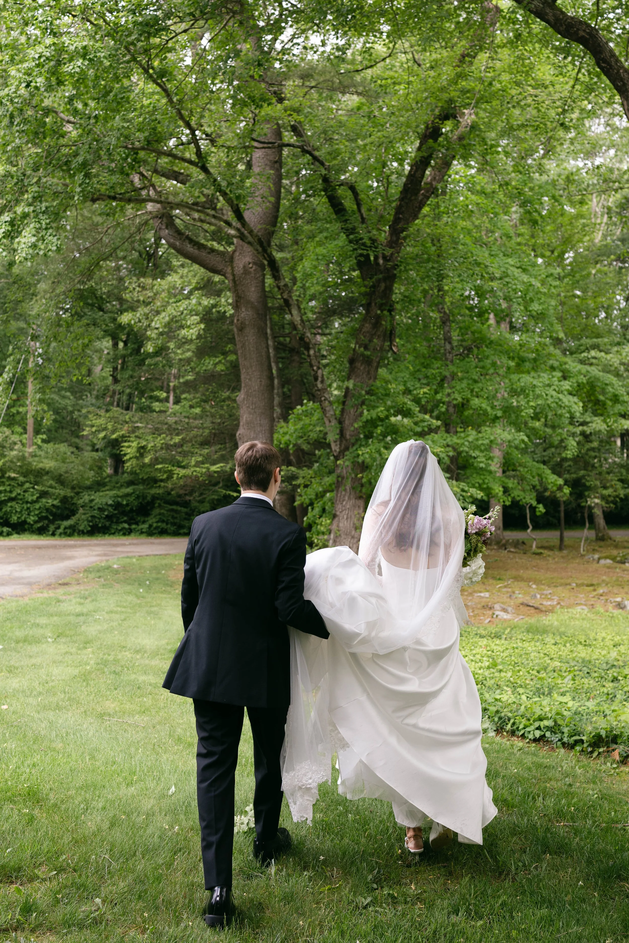 A bride in a white wedding dress and veil walking with a groom in a black suit through a grassy park with large trees.