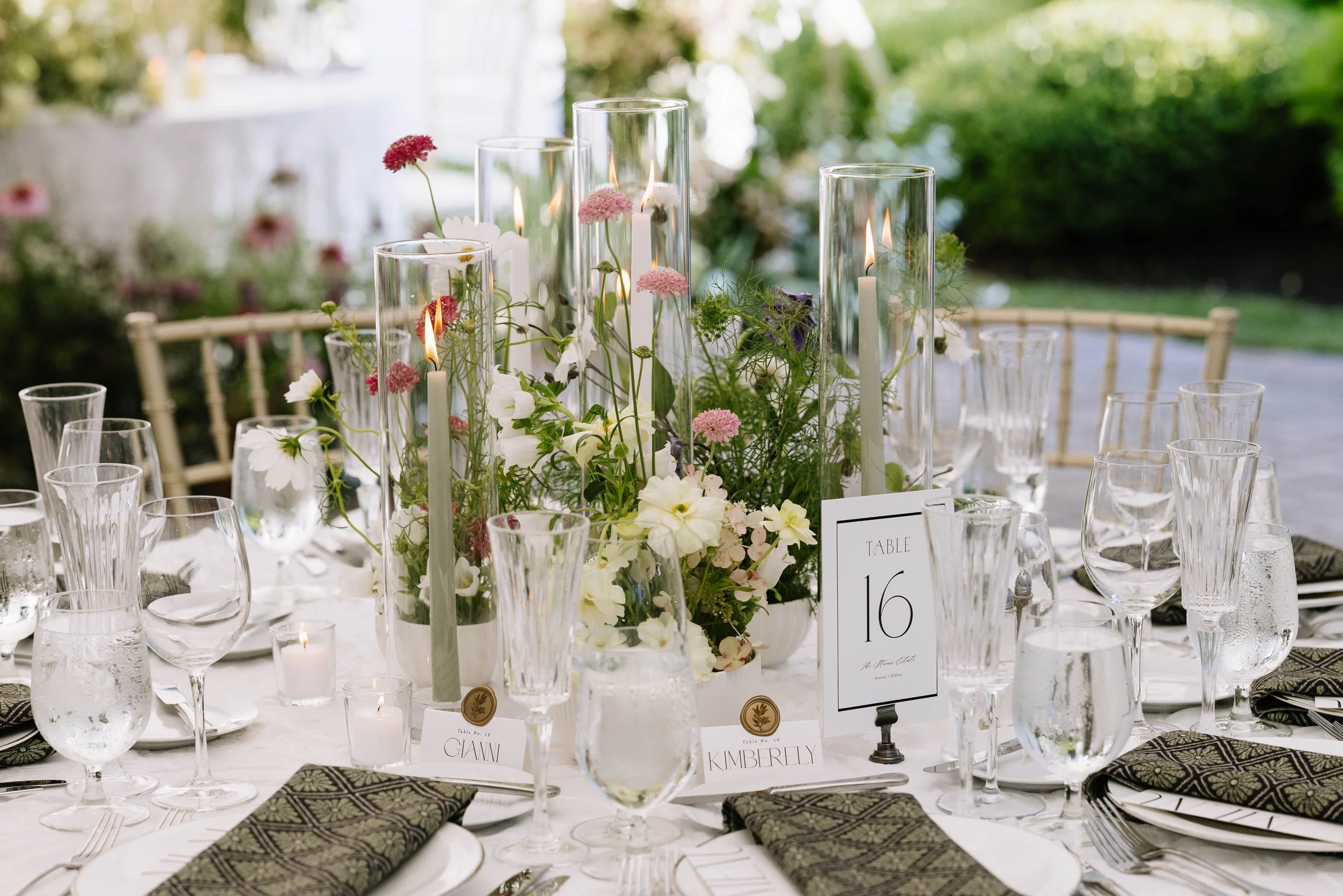 Elegant outdoor banquet table with floral centerpiece, candles, glasses, black napkins, and table number sign.