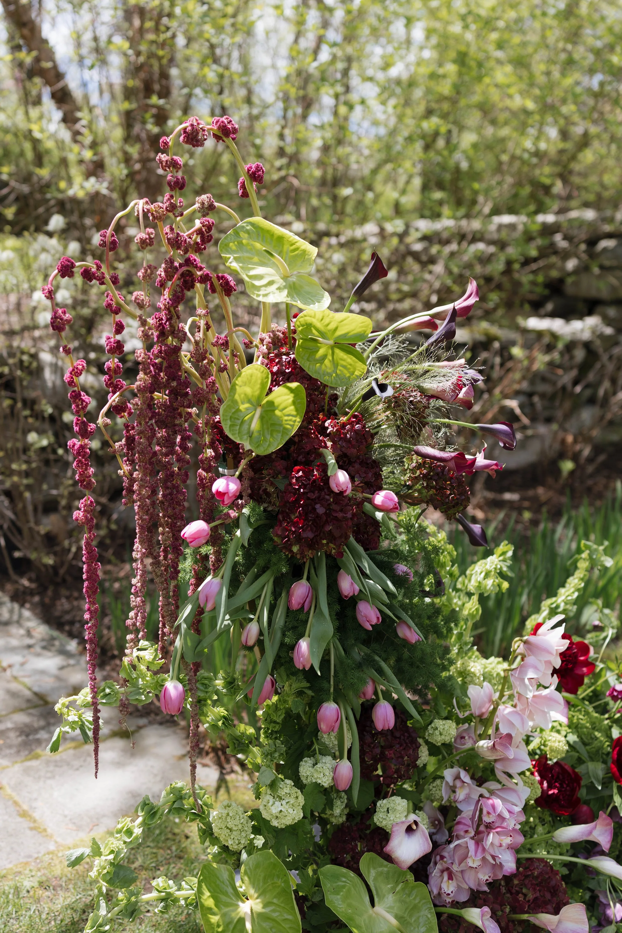 A colorful flower arrangement with purple, pink, white, and green flowers, including calla lilies, hydrangeas, and other assorted flowers, set outdoors in a garden.