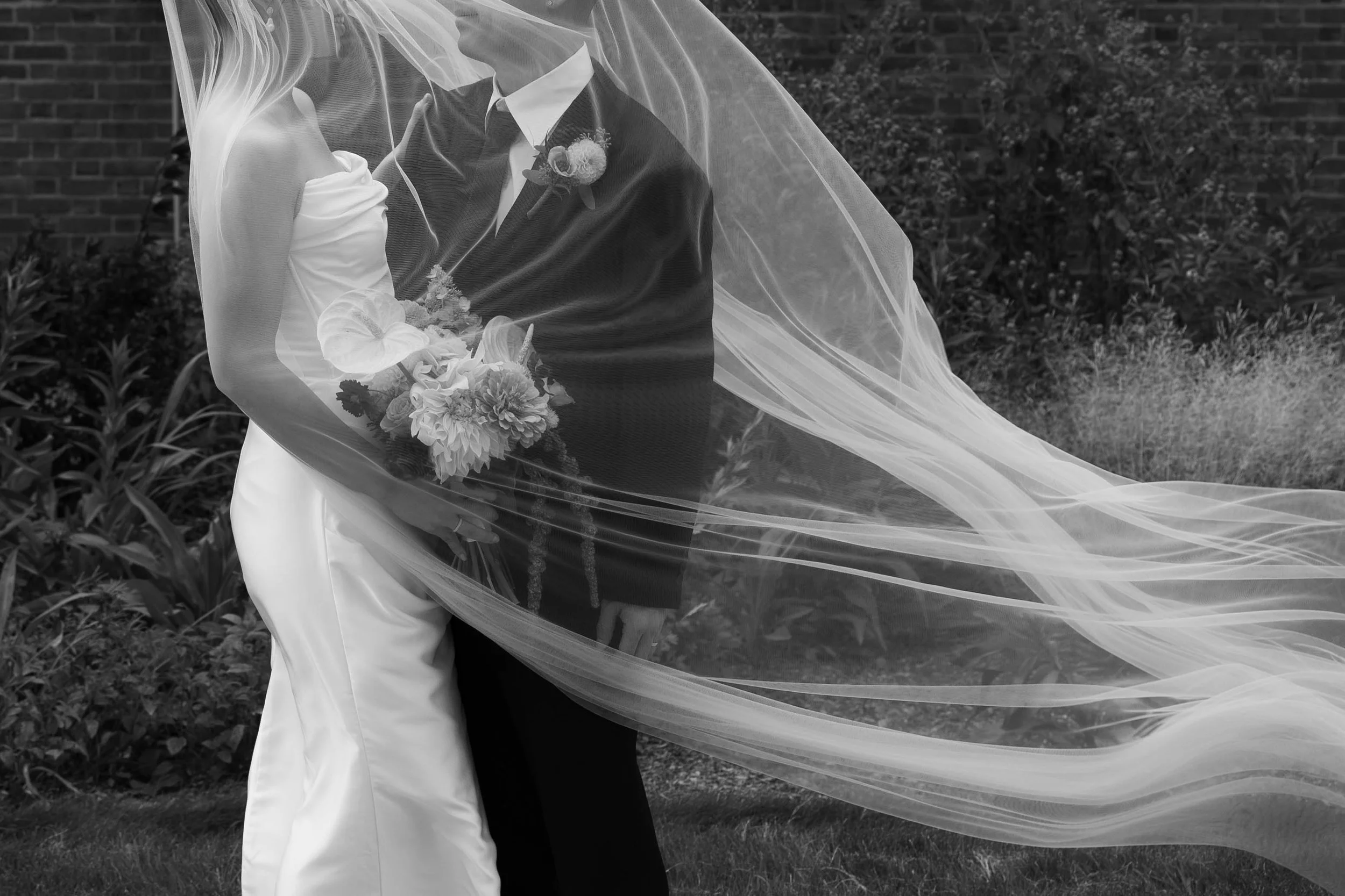 A bride holding a bouquet of flowers standing close to a groom, with their faces not visible, outdoors with a brick wall and plants in the background. The bride's veil is flowing in the wind.