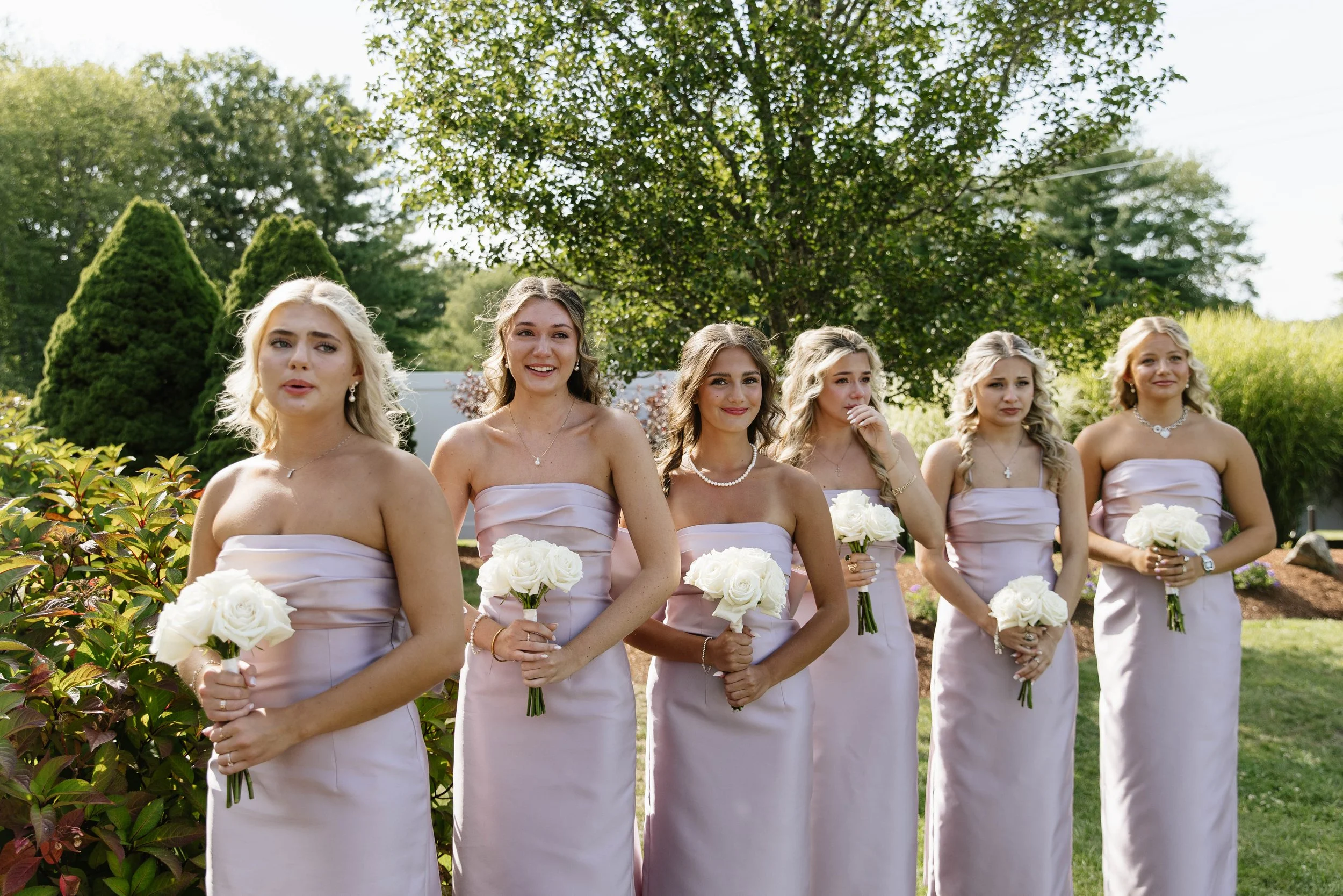 Six bridesmaids standing outdoors in a garden during daytime, wearing lavender strapless dresses, holding white roses, and smiling.