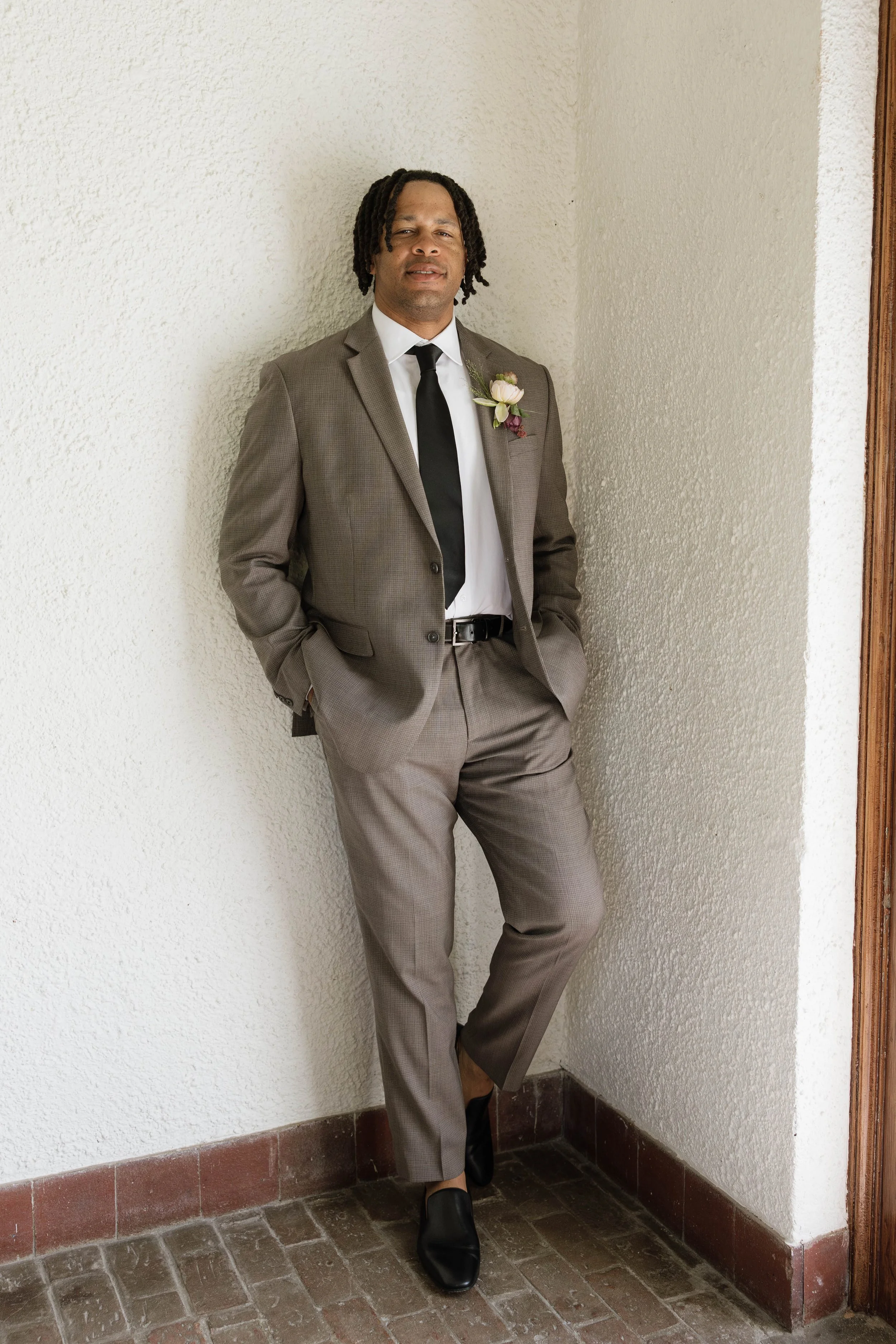 Man in a gray suit with a white shirt and black tie, leaning against a corner with his hands in his pockets, wearing a boutonnière, standing on a tiled floor next to a white stucco wall.
