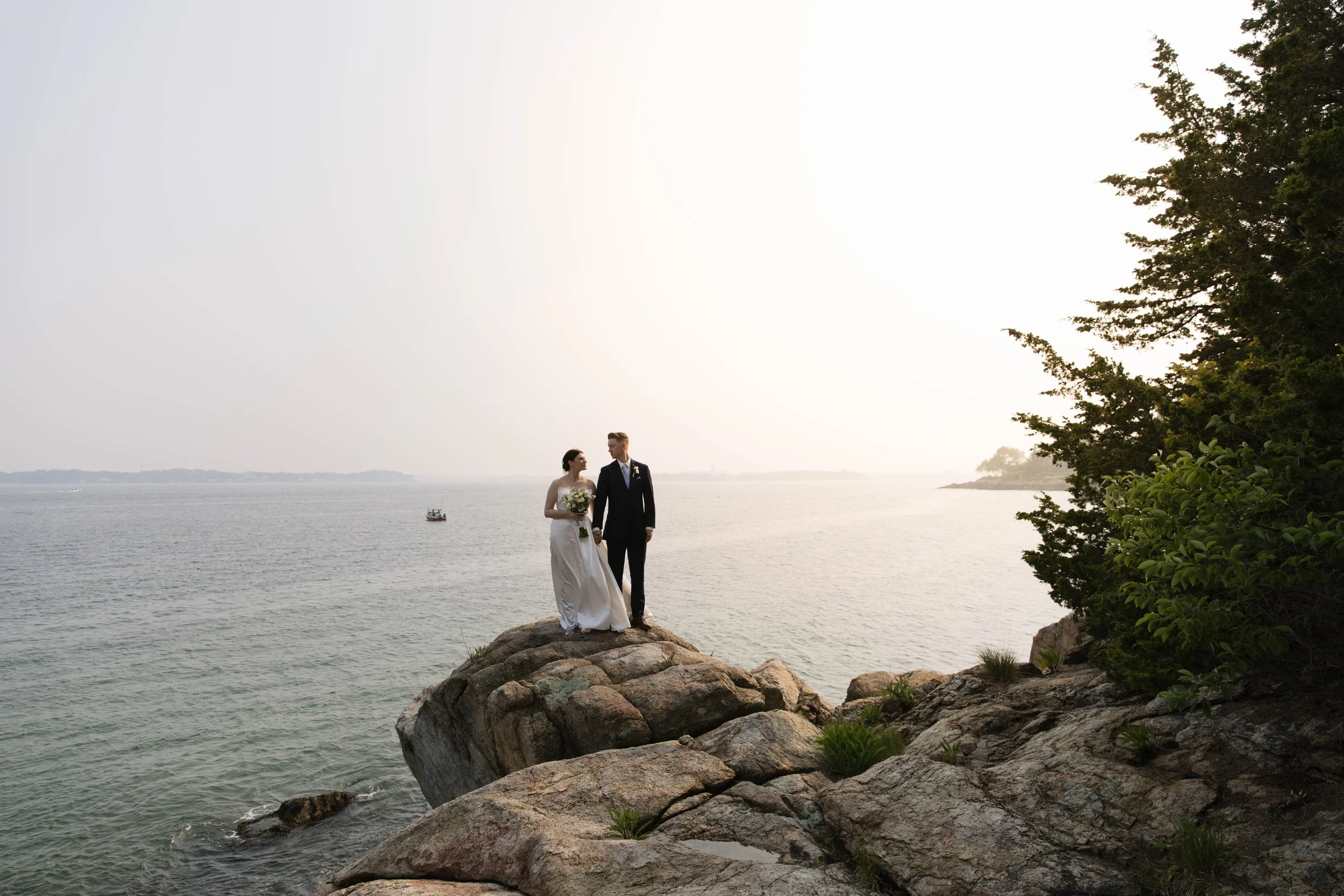 A bride and groom standing on rocks by the water, holding hands, with a boat in the distance and trees on the side.