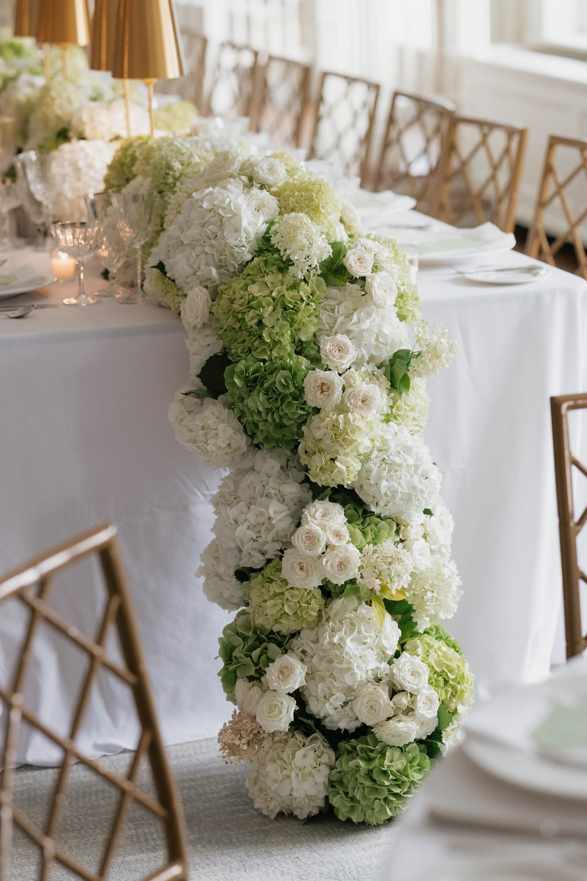 Elegant wedding table with flower arrangement in white, cream, and green, draped on a white tablecloth, with chairs, plates, and candles.