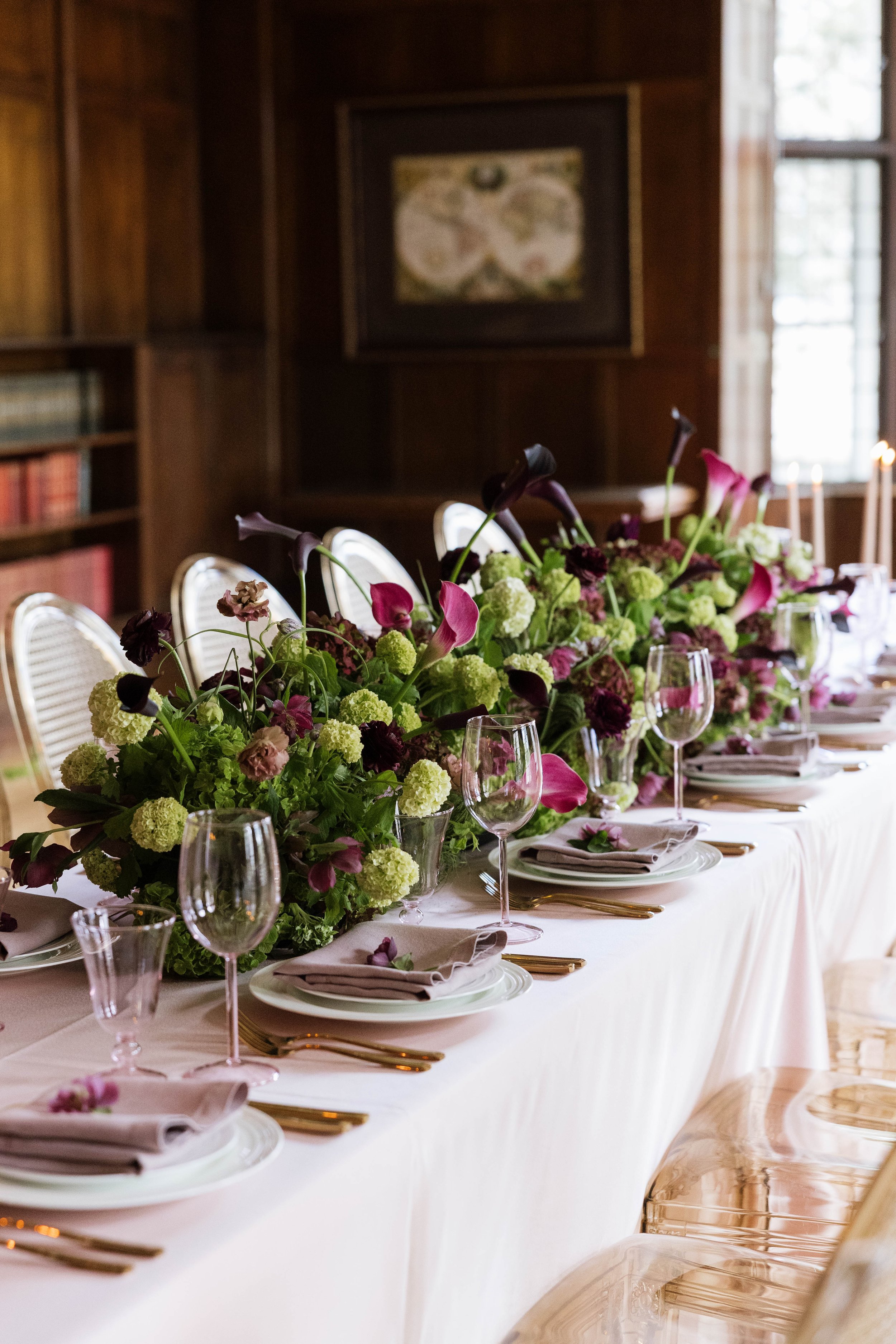 An elegant dining table set for a formal event, decorated with a large floral centerpiece including pink calla lilies and green hydrangeas, surrounded by wine glasses, plates, and beige napkins with gold cutlery, in a room with wooden walls and large