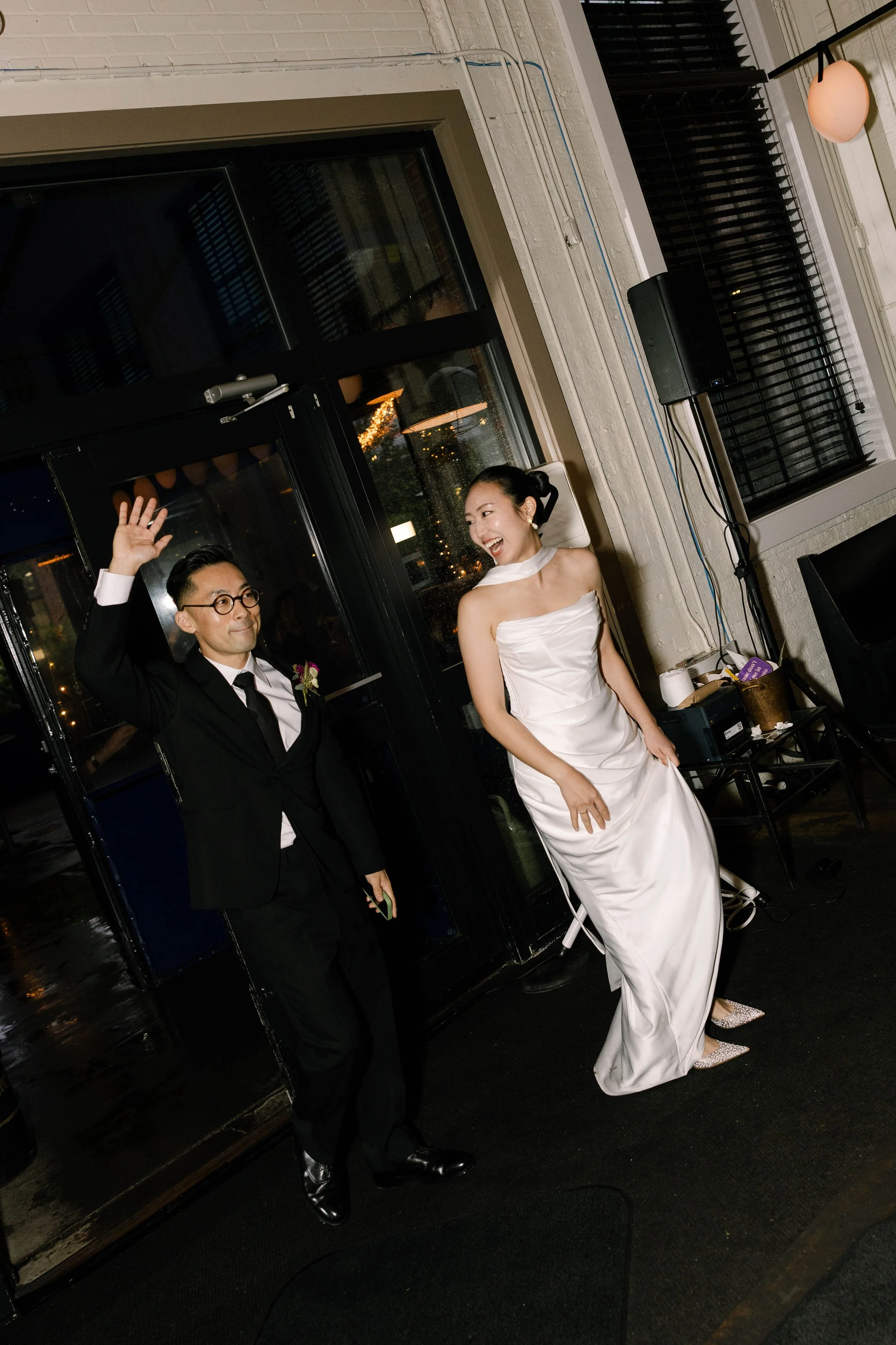 A couple dressed in wedding attire, with the groom in a black tuxedo and the bride in a white wedding gown, smiling and waving at a celebration or reception.