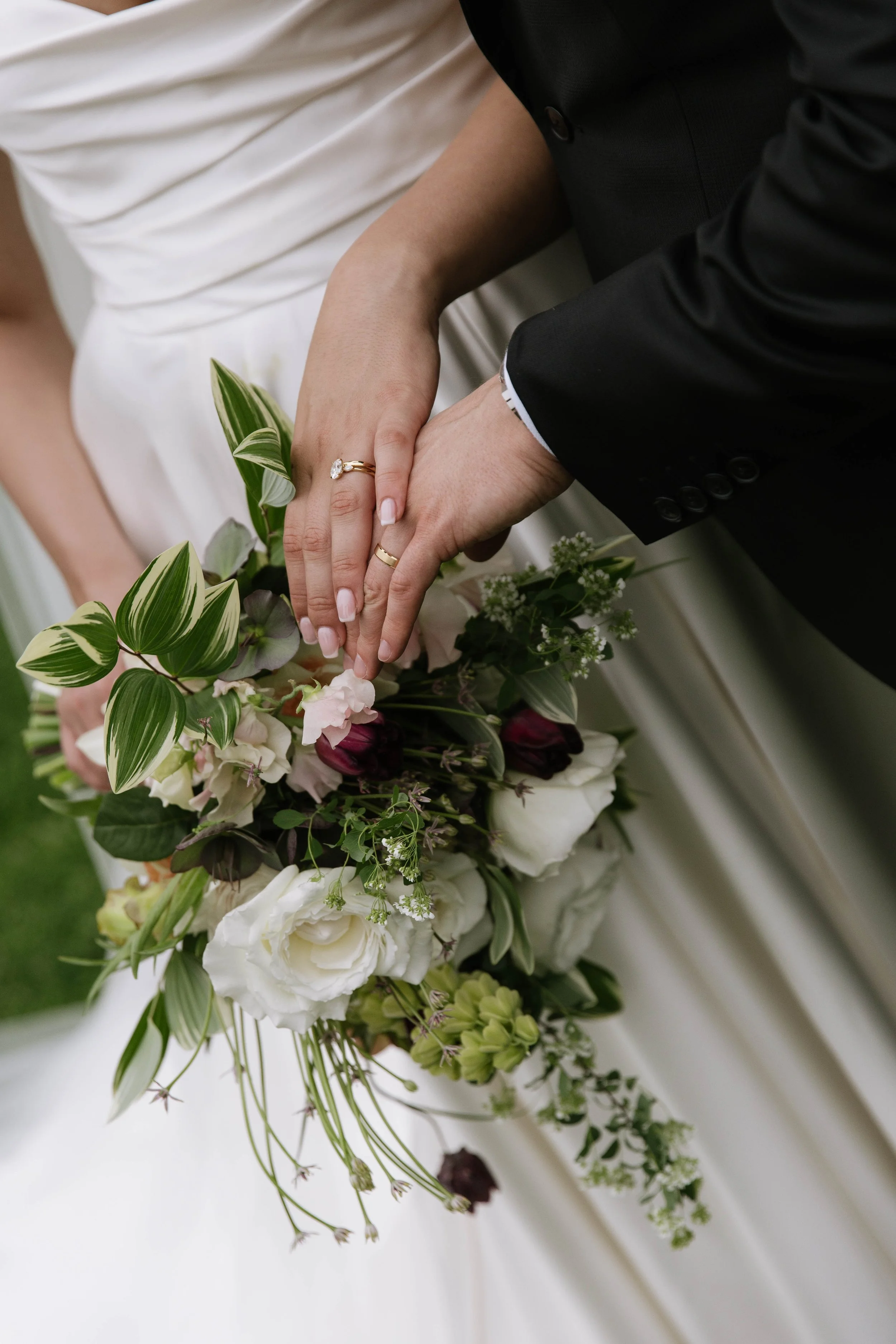 Close-up of a bride and groom holding hands over a bridal bouquet, showing wedding rings and wedding attire.