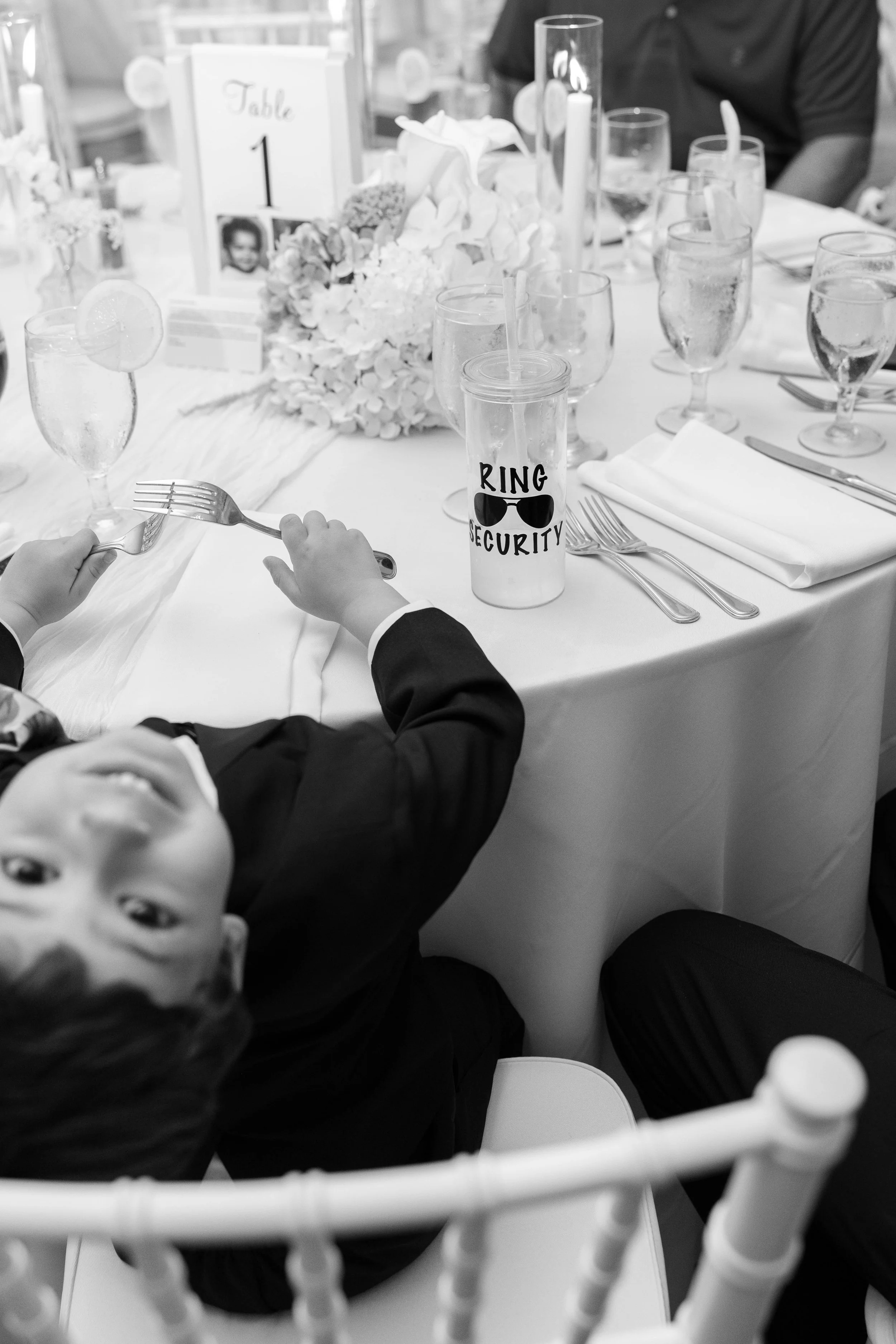 Child in formal attire sitting at a decorated table with drinks and floral centerpieces at a celebration, with a sign that says 'Ring Security'.