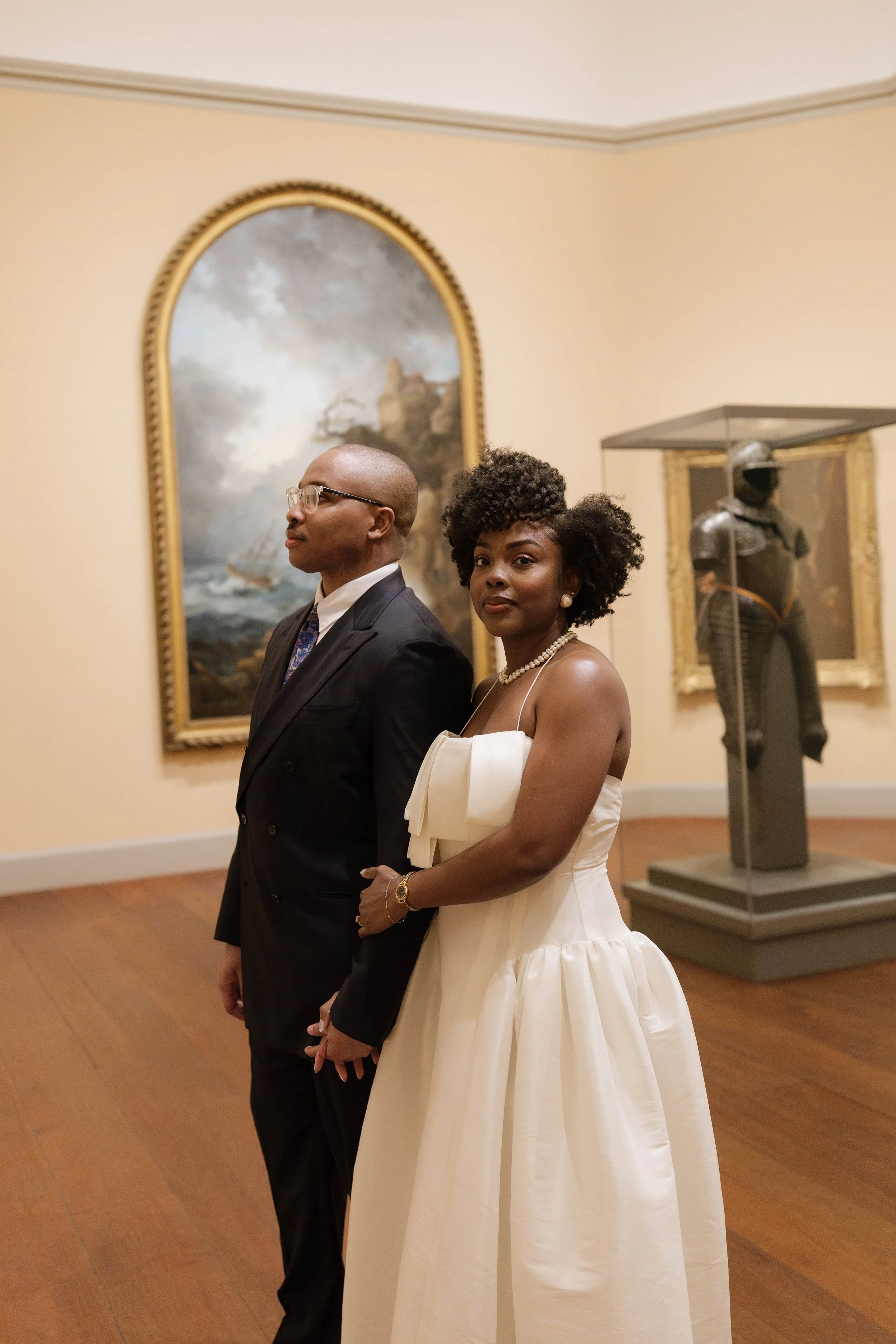 A man and woman stand in an art gallery, dressed formally. The man wears a suit and glasses, and the woman wears a white strapless dress and pearl jewelry. Behind them are paintings and sculptures displayed in the gallery.