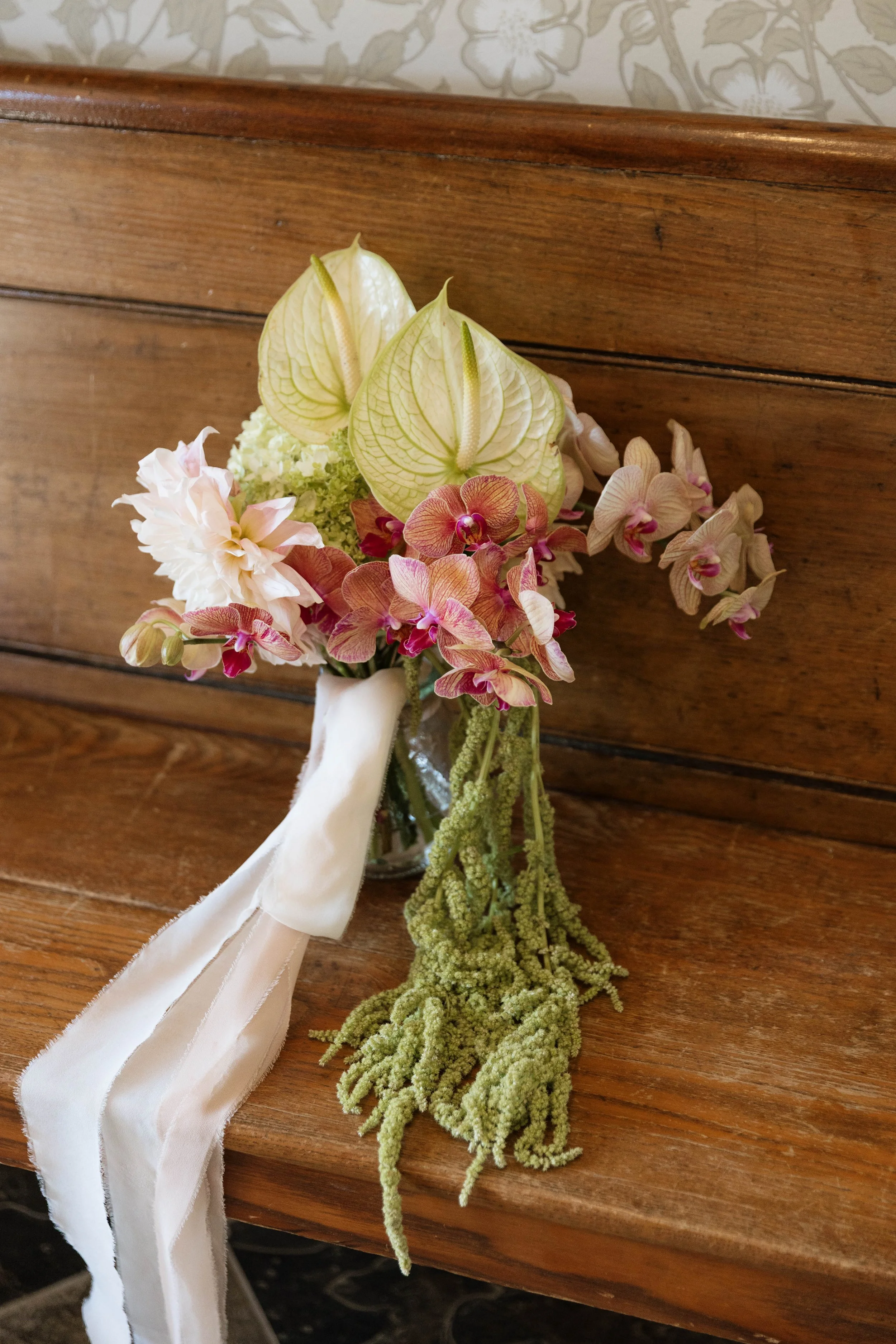 A floral arrangement with anthurium, orchids, and a hanging green plant in a glass vase, placed on a wooden surface.