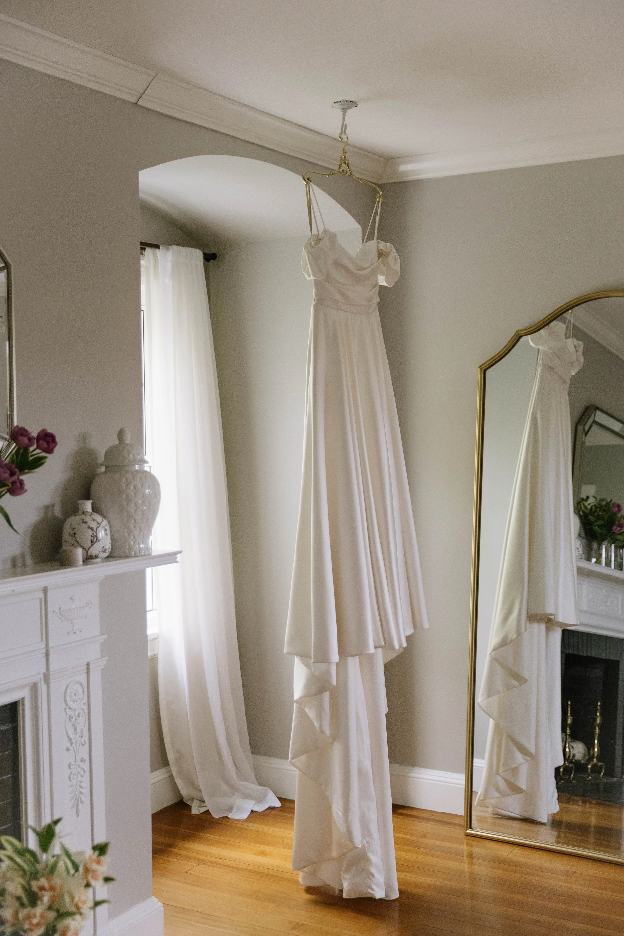 A white wedding dress hanging from the ceiling in a softly lit room, with a mirror reflecting the dress and a window with white curtains.