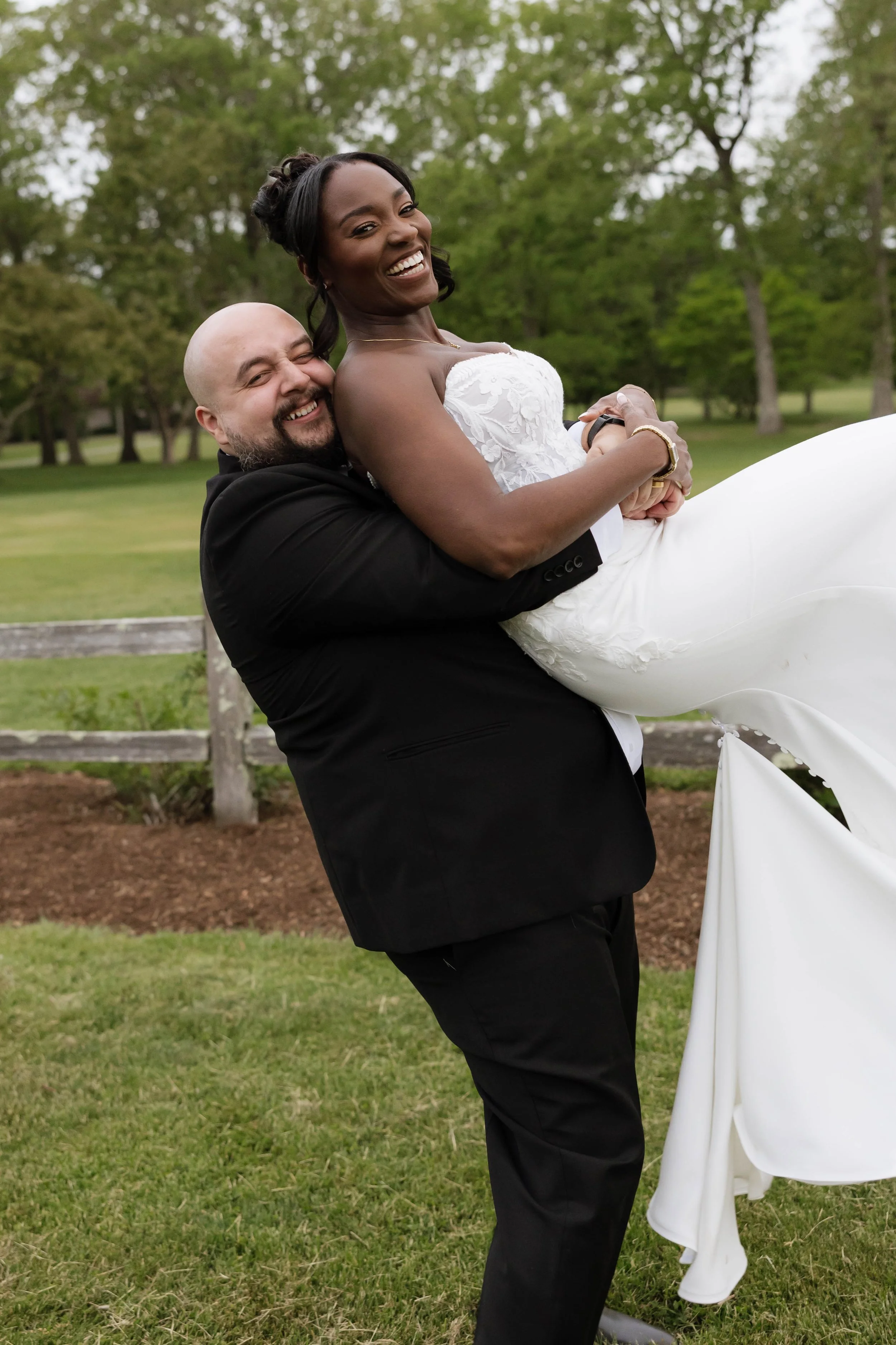 A smiling man holding a smiling woman in a white wedding dress outdoors with green trees in the background.