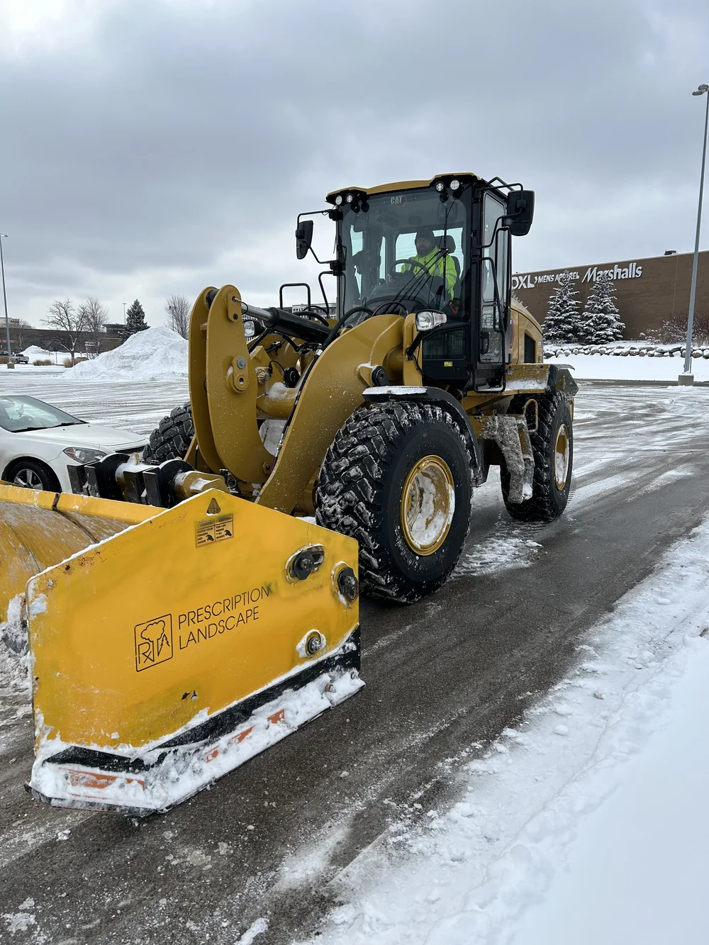 A yellow snowplow clearing snow from a parking lot under cloudy sky. A man in a high-visibility jacket is inside the snowplow's cab. A shopping mall called Marshalls is visible in the background.