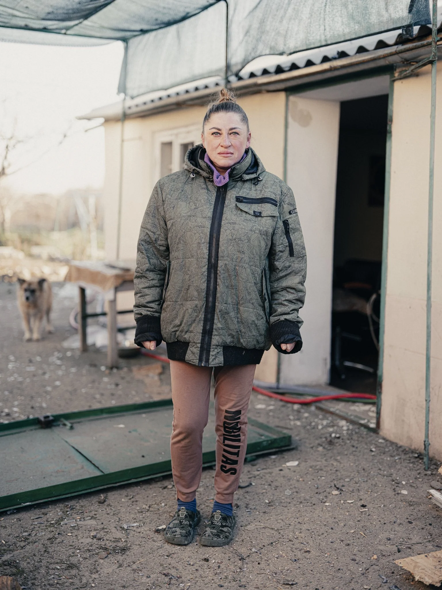 A woman stands outdoors in front of a small building or shed, wearing a gray patterned jacket and pink pants with 'SQUAD' written on the side, with a neutral expression on her face. A dog is visible in the background near a table.