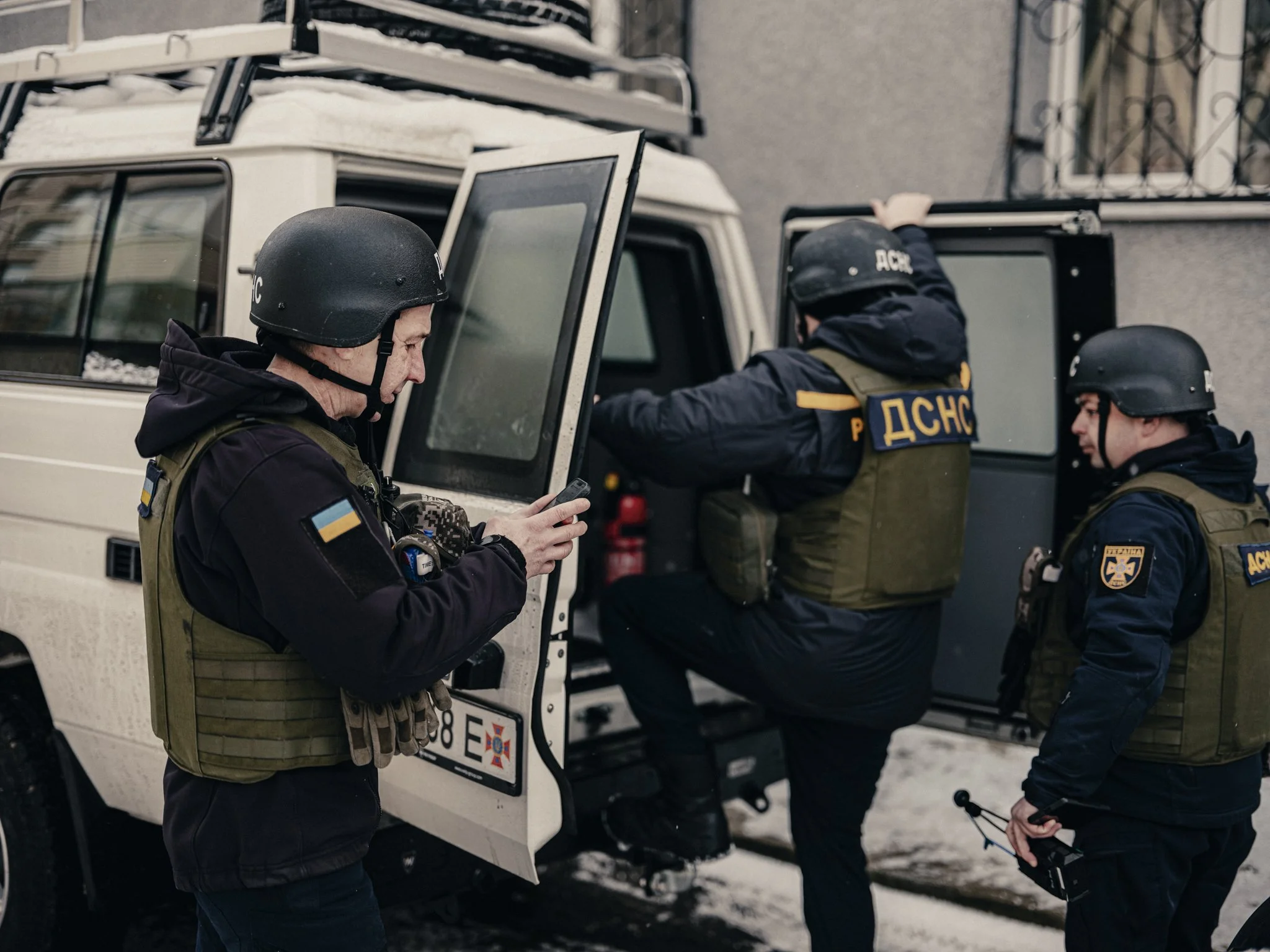 Three police officers wearing black helmets and tactical vests with Ukrainian flags, conducting an operation outside a building in winter. One officer is using a cellphone, another is climbing into a vehicle, and the third is holding equipment.