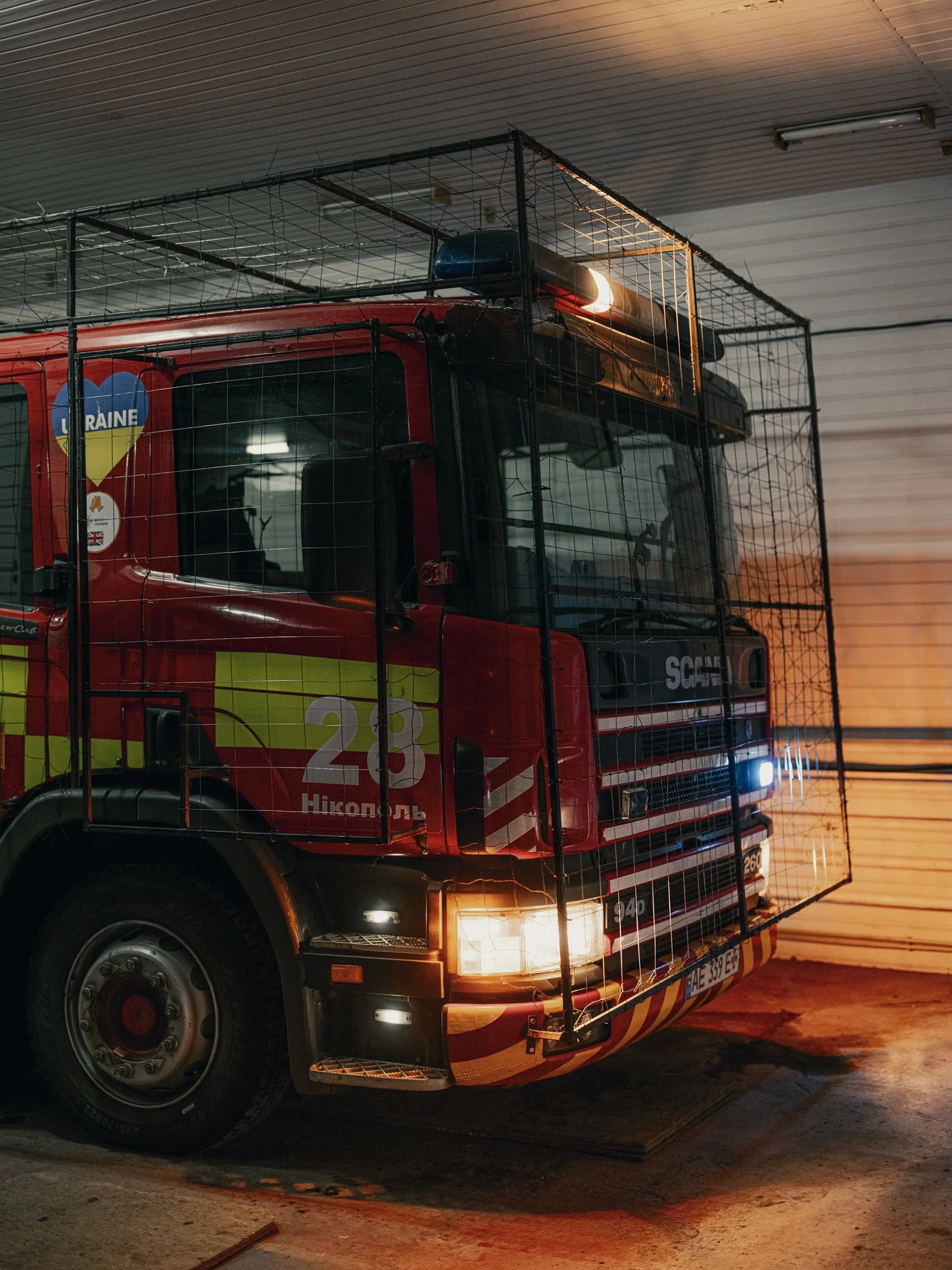 A red fire truck with a metal cage structure attached to the front, parked inside a garage, with lights on.