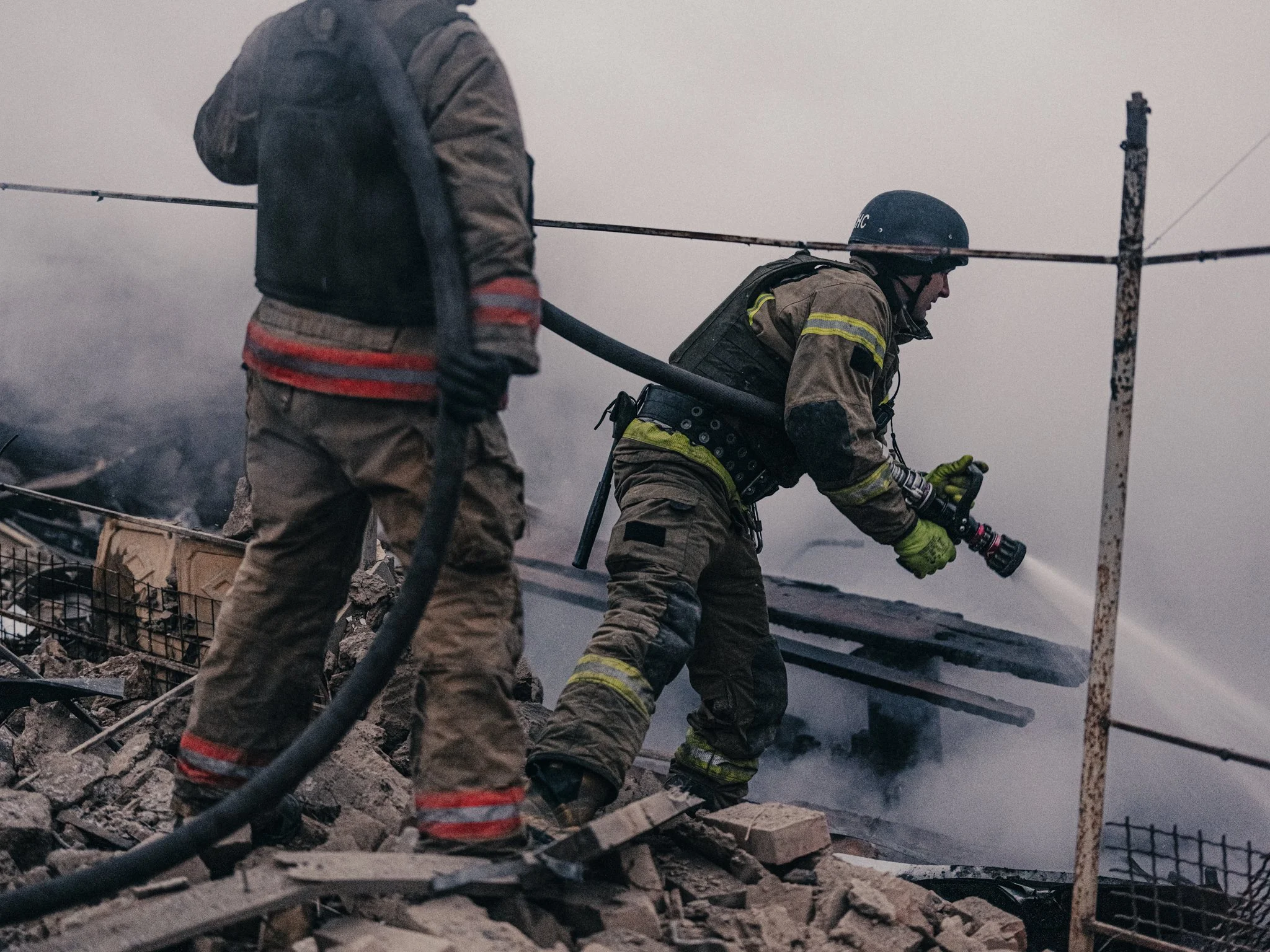 Two firefighters in full gear working amid debris and smoke, one holding a hose, during a firefighting effort on a damaged building.
