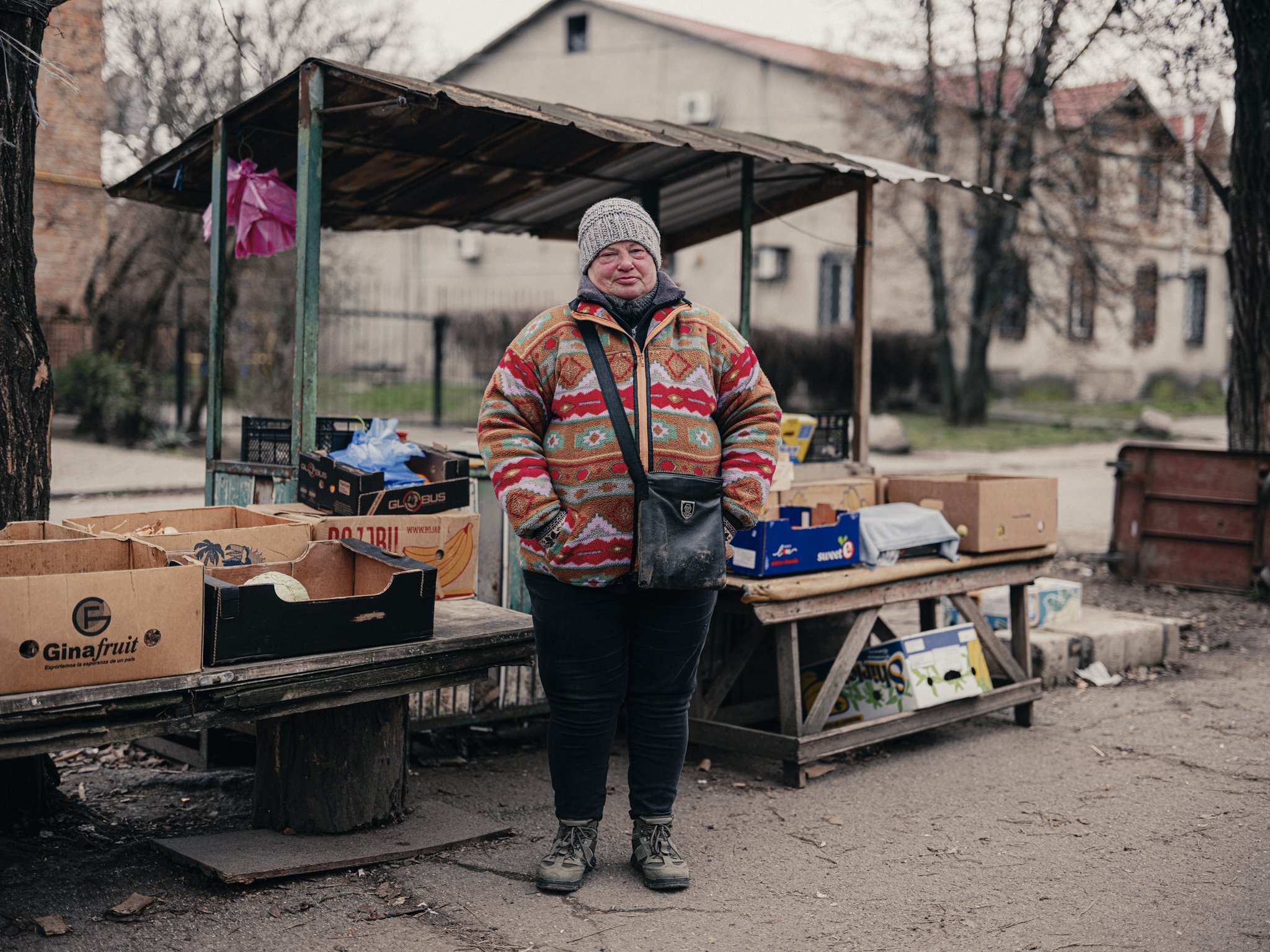 A woman standing at an outdoor market stall, wearing a colorful sweater, gray beanie, and gray shoes, with boxes of produce and other goods on wooden tables, in an urban setting with trees and buildings in the background.