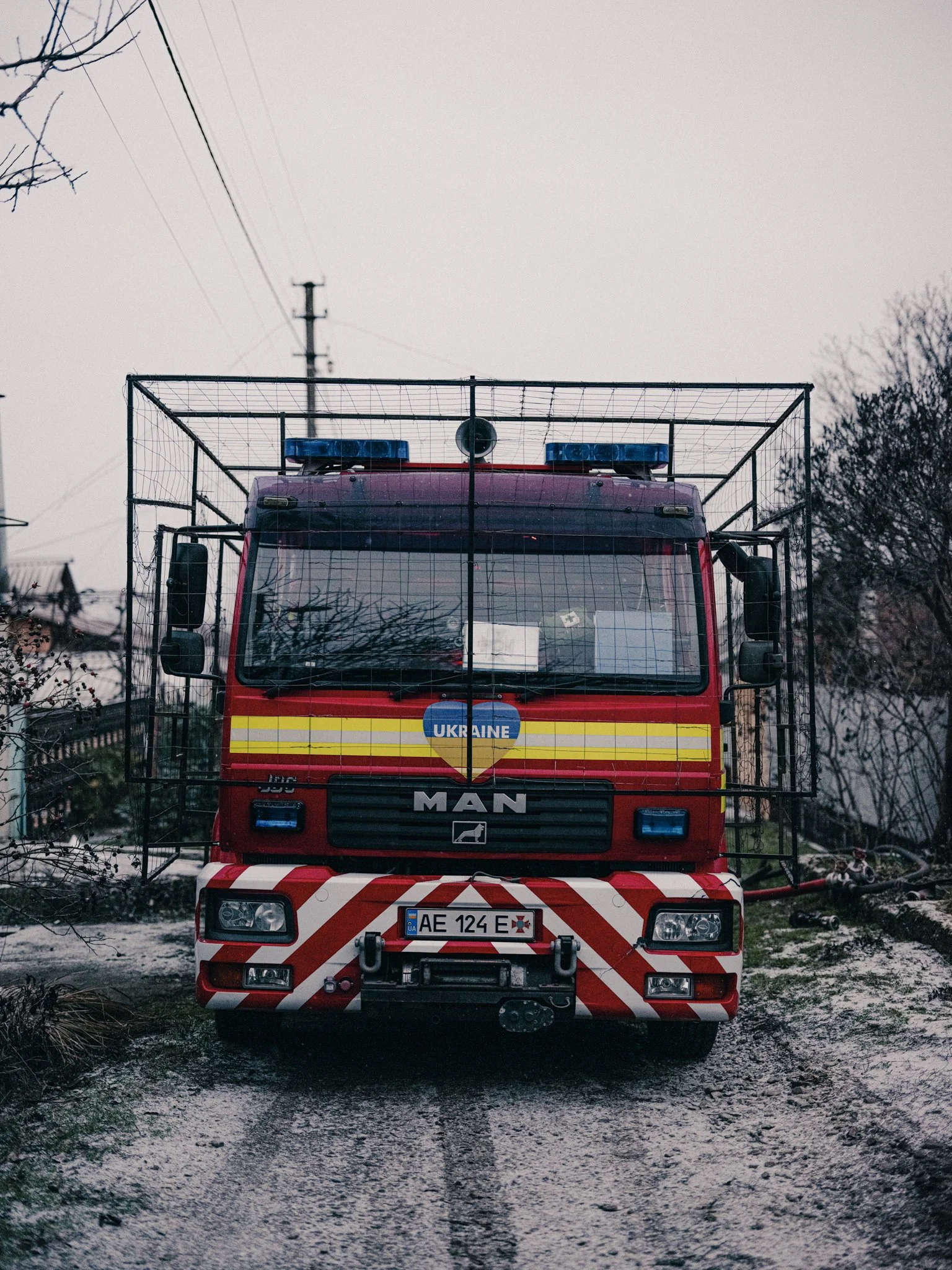 Front view of a red fire truck with a metal cage around the top, parked on a dirt road, with a cloudy sky and power lines overhead.