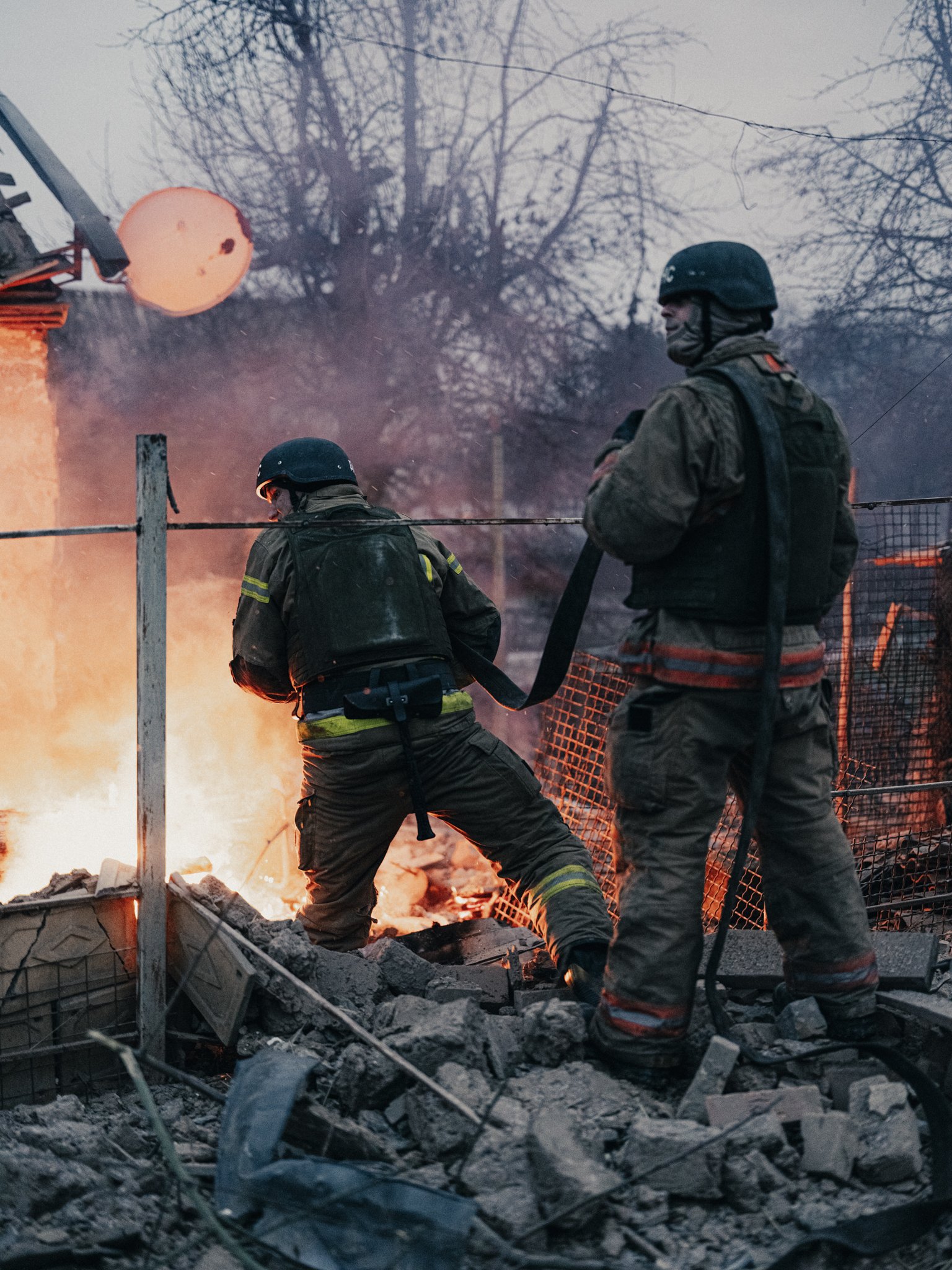 Two firefighters battling a fire amidst debris and rubble outside, with trees in the background and a large light fixture overhead.