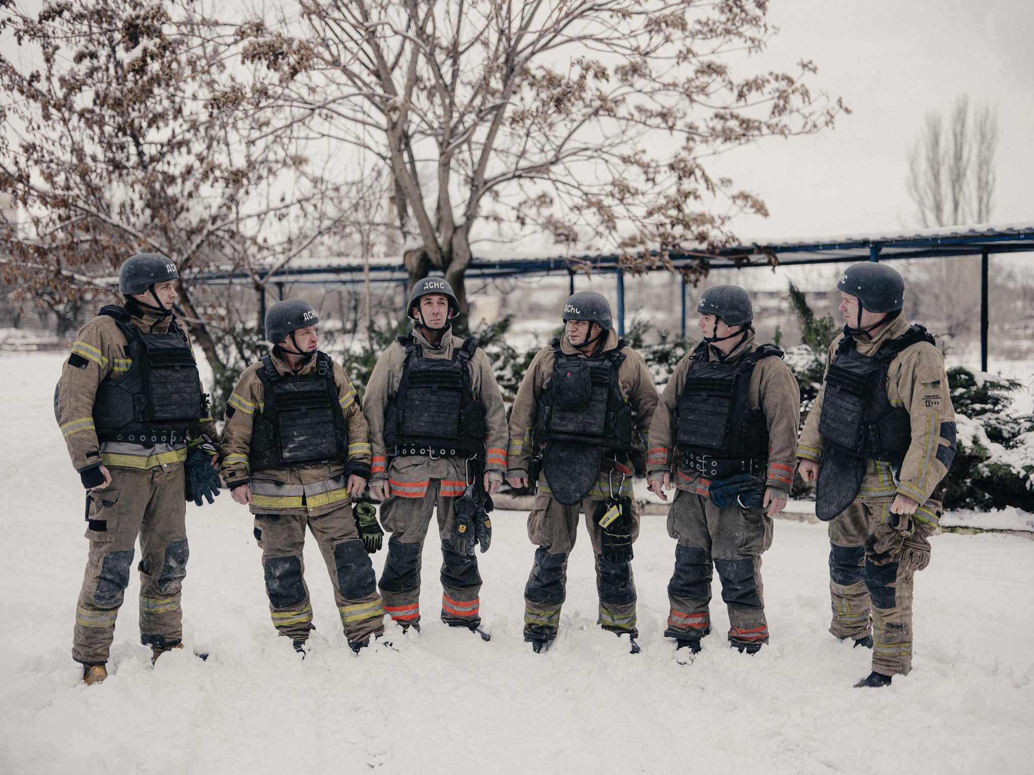 Group of six firefighters standing in snow, wearing helmets and protective gear, with trees and a fence in the background.