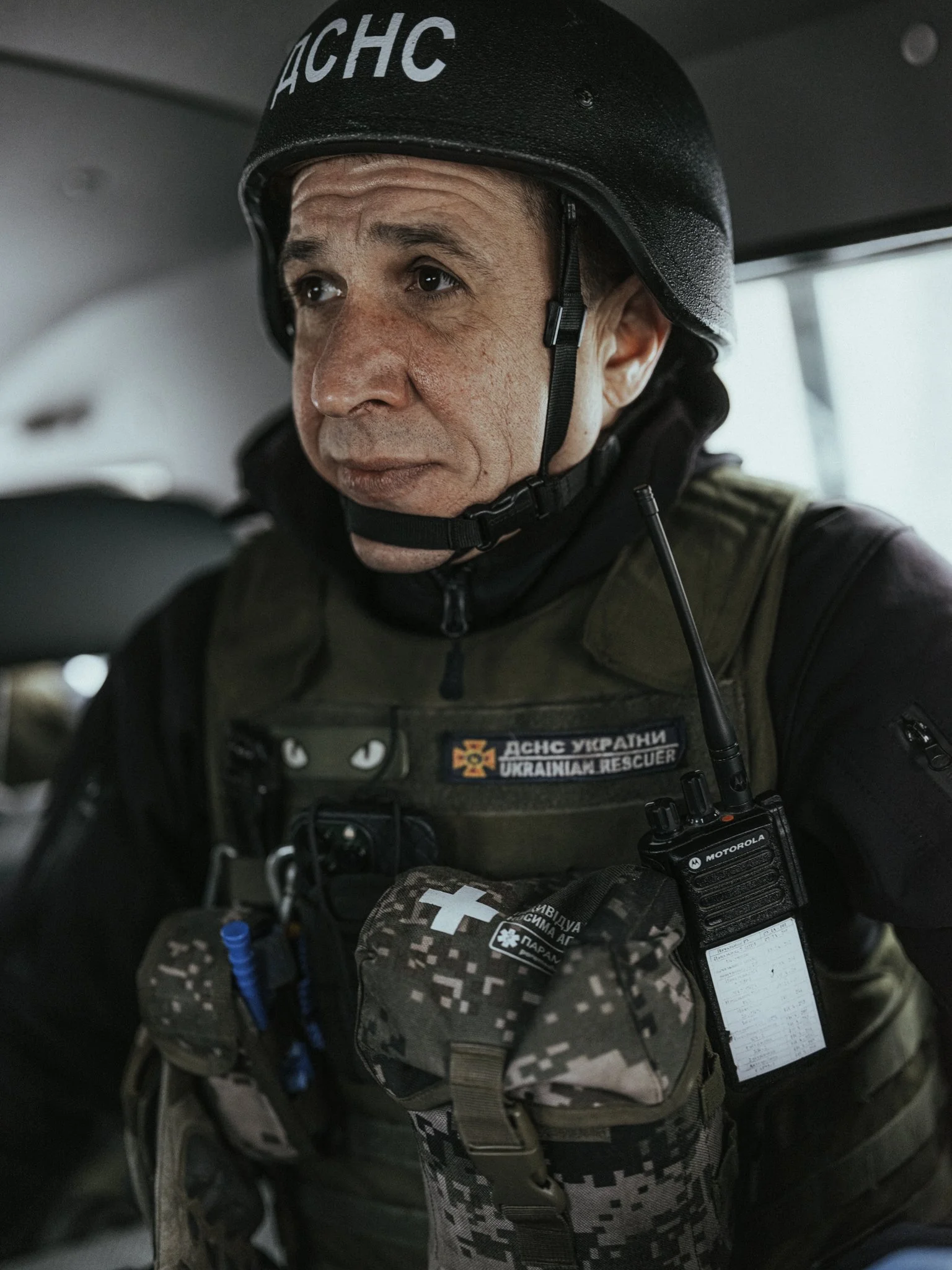 A Ukrainian rescue worker wearing a black helmet and tactical gear inside a vehicle.