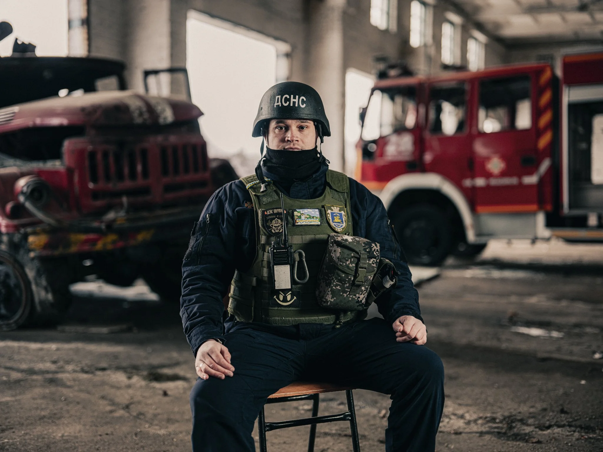 A firefighter sitting on a chair inside a building with firefighting vehicles in the background, wearing a helmet and protective gear.