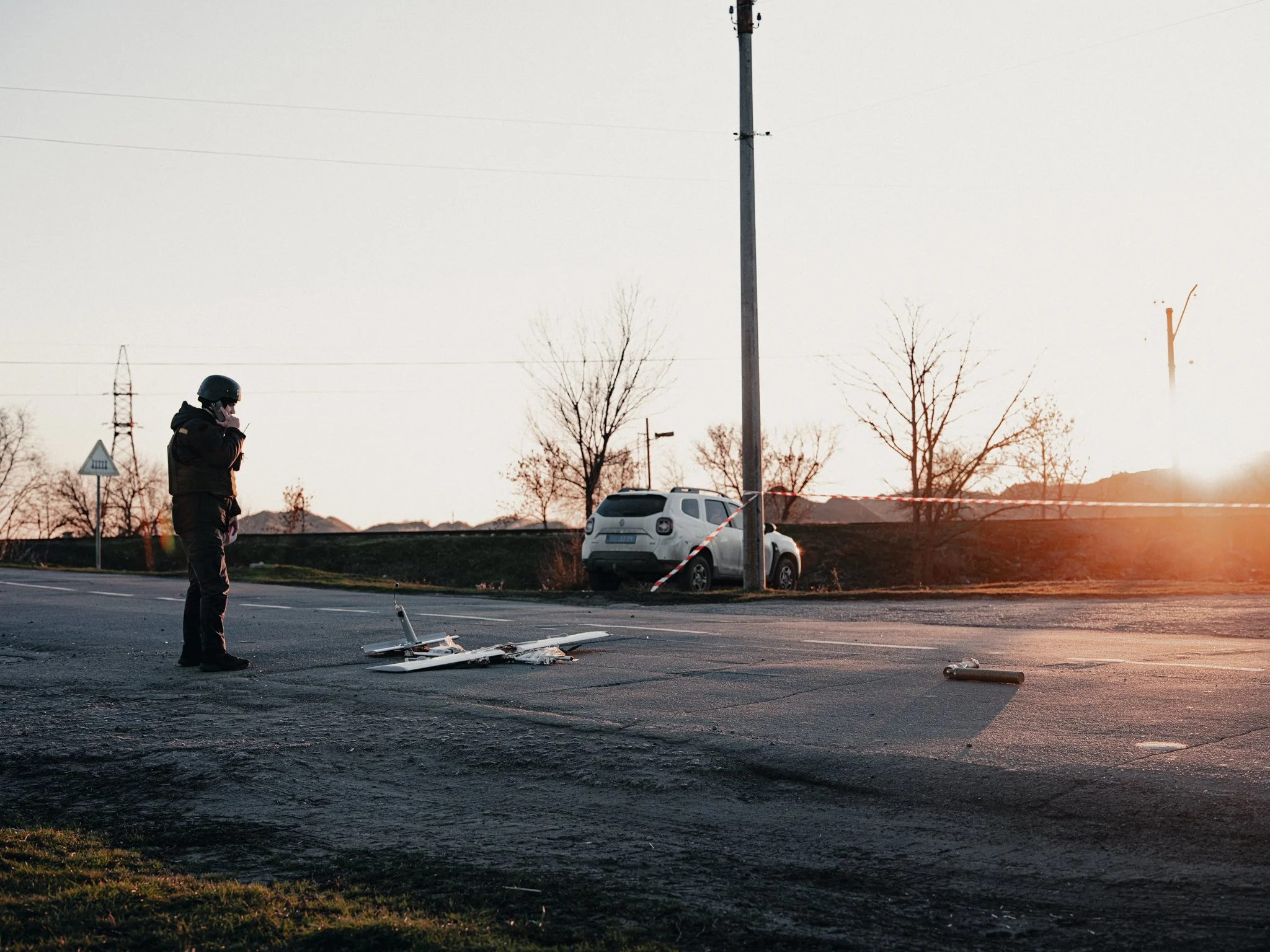 Scene of a traffic accident with a fallen drone on the road, a police officer speaking on a phone, and a white car in the background near a utility pole, at sunset.