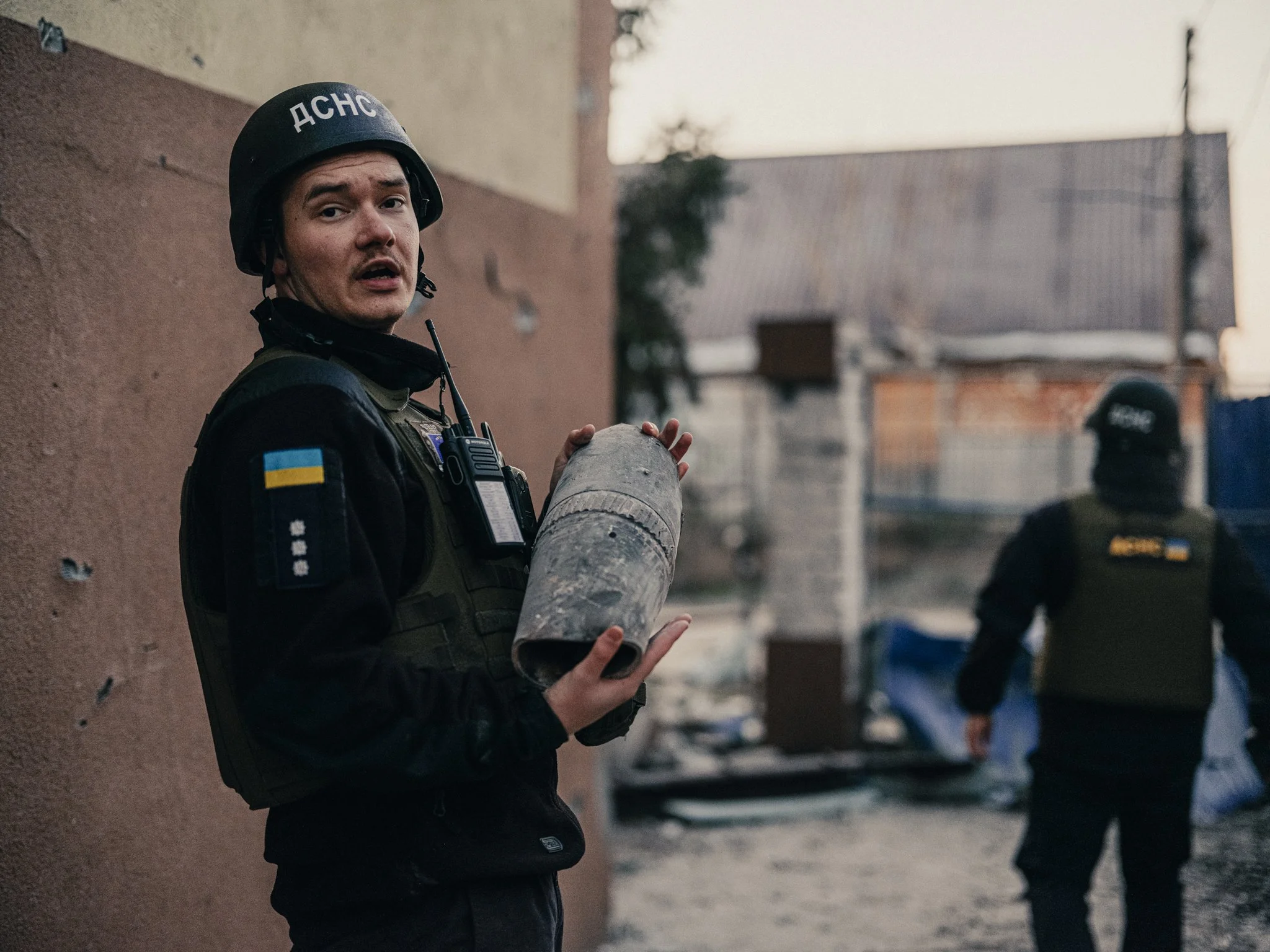 A police officer in tactical gear holding a tear gas canister standing outside, with another officer in the background during what appears to be a protest or conflict scene.