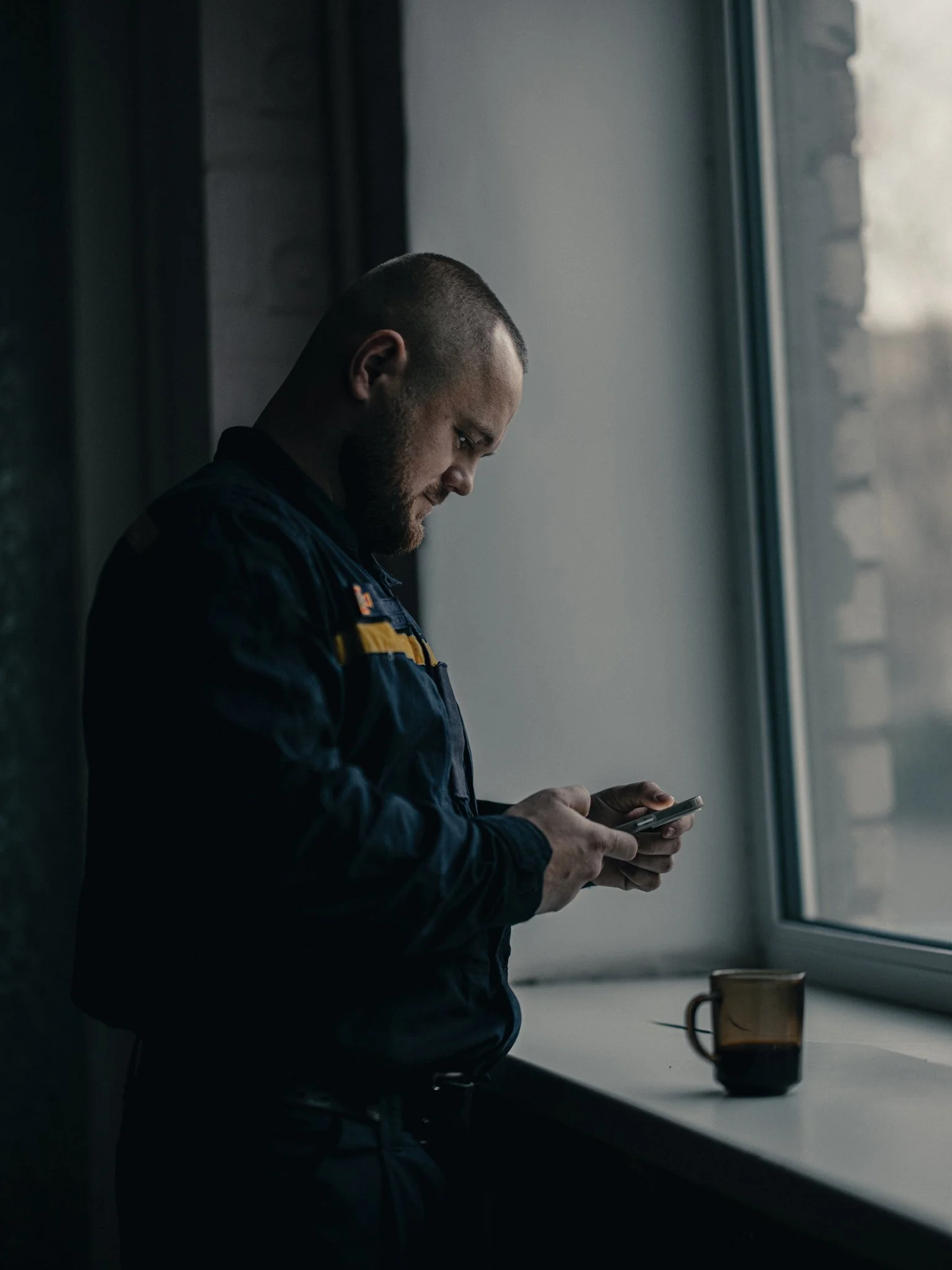 A man in a dark uniform stands by a window, looking at his phone with a mug on the windowsill.