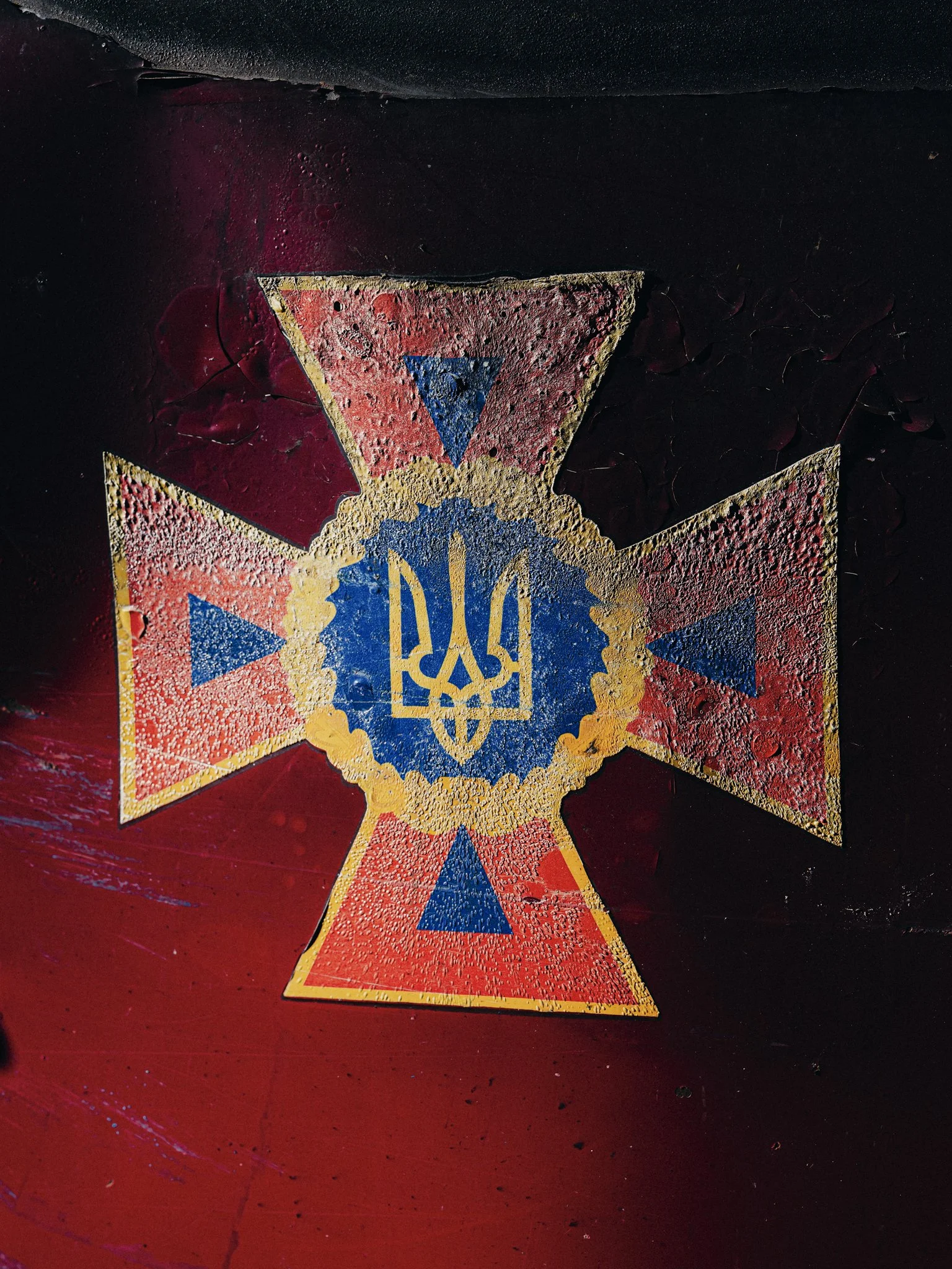 Close-up of a red and black insignia with a central blue and yellow emblem featuring a trident symbol, surrounded by a yellow border and triangular shapes.