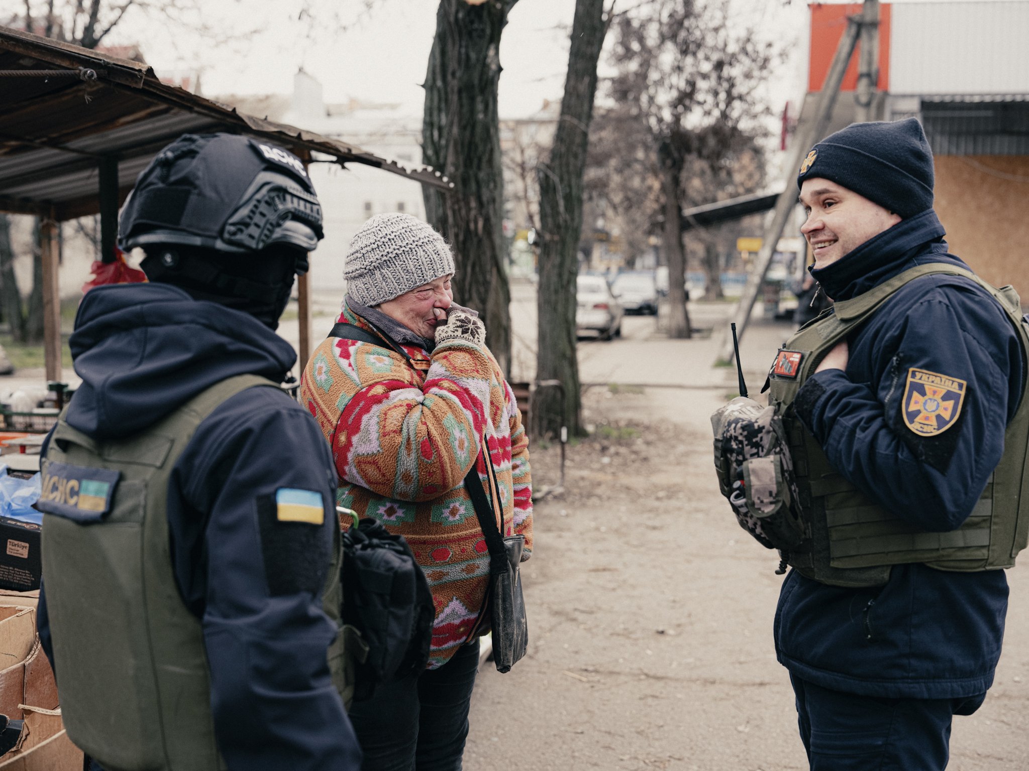 The woman is wearing a colorful sweater, gray knit hat, and gloves, and is covering her nose with her hand. The officers are in uniform with Ukrainian patches and badges, smiling and engaging in conversation. The background shows trees, parked cars, 