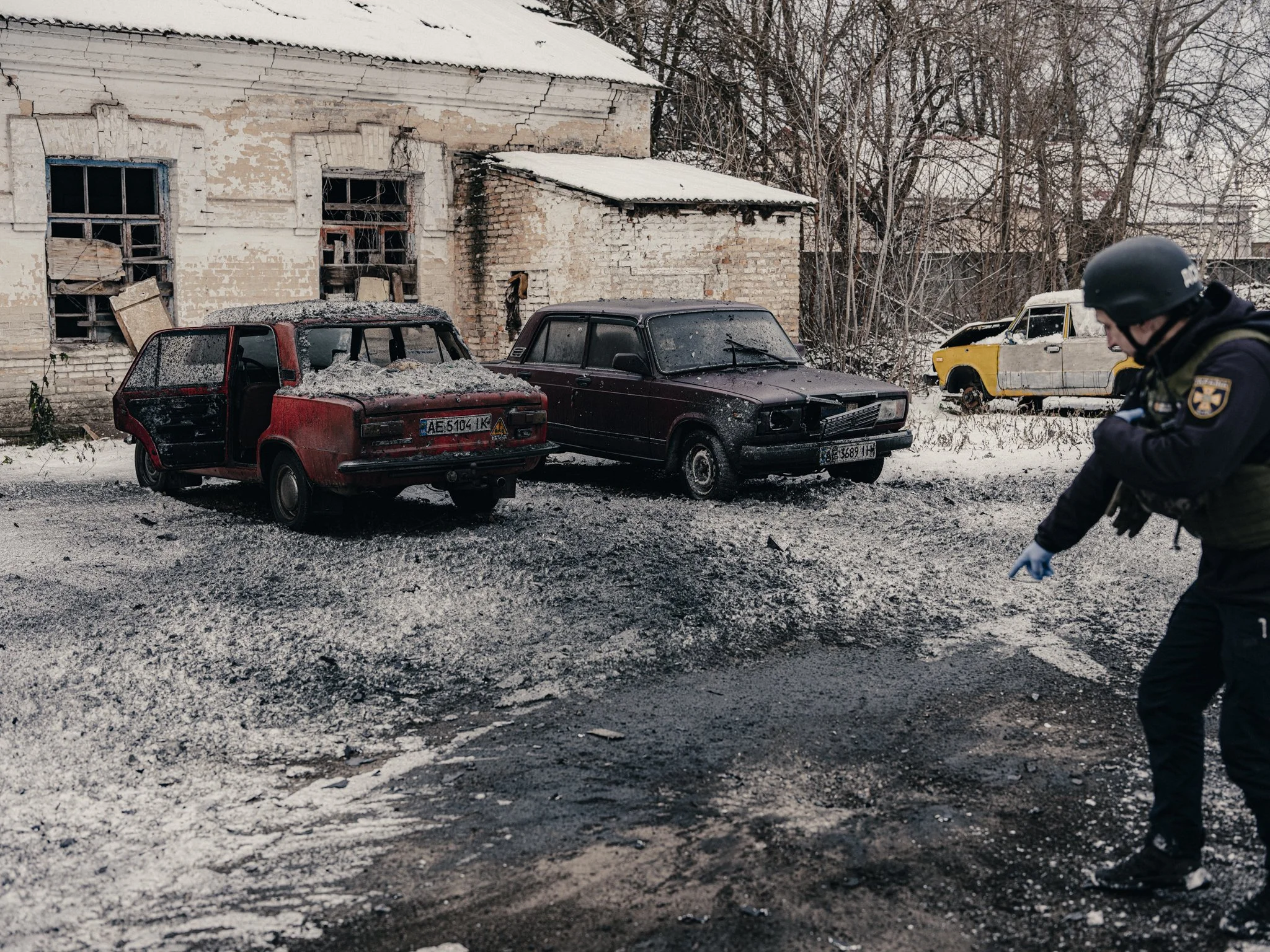 A police officer pointing at burned out cars with damaged and broken windows, parked in front of an old, deteriorated brick building in a winter setting with snow on the ground.
