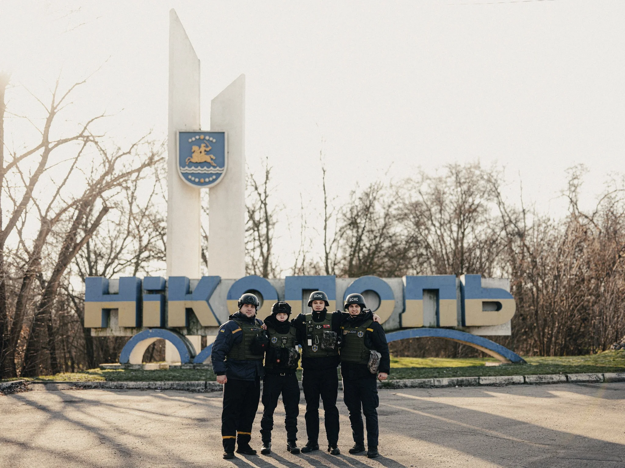 Four law enforcement officers standing together in front of the entrance sign to Nikopol, Ukraine, with leafless trees in the background.