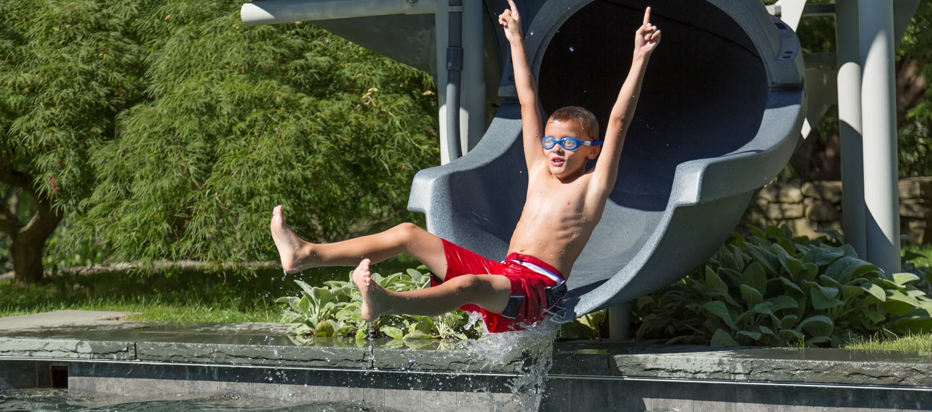A young boys comes down a slide into his backyard pool.