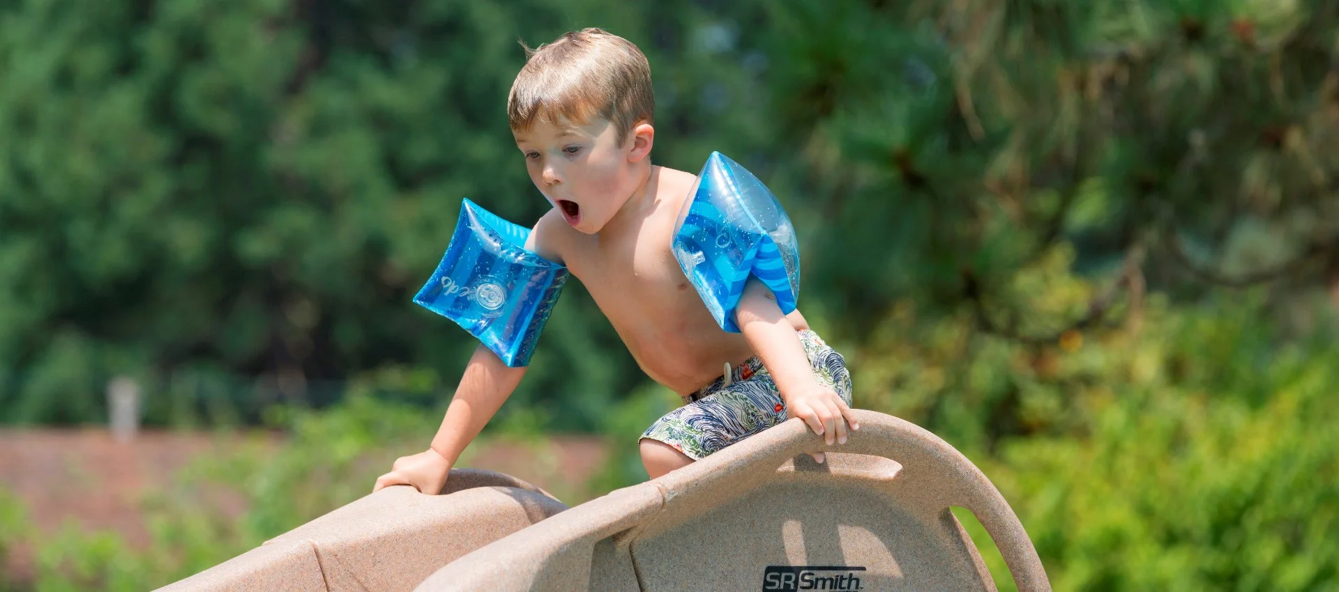 A young boy wearing arm float gets ready to go down a slide in his family’s backyard pool.