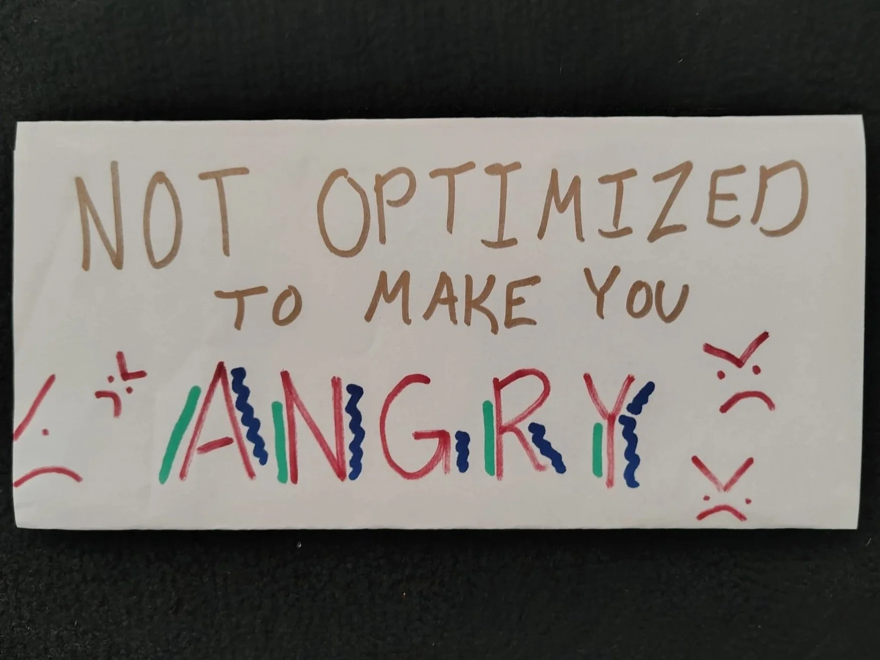 Handwritten graphic on a folded piece of A4. In bronze Sharpie, the text: "NOT OPTIMIZED TO MAKE YOU" and in red Sharpie, "ANGRY". Several icons related to anger (jagged lines, a few ">:(" symbols) accompany.