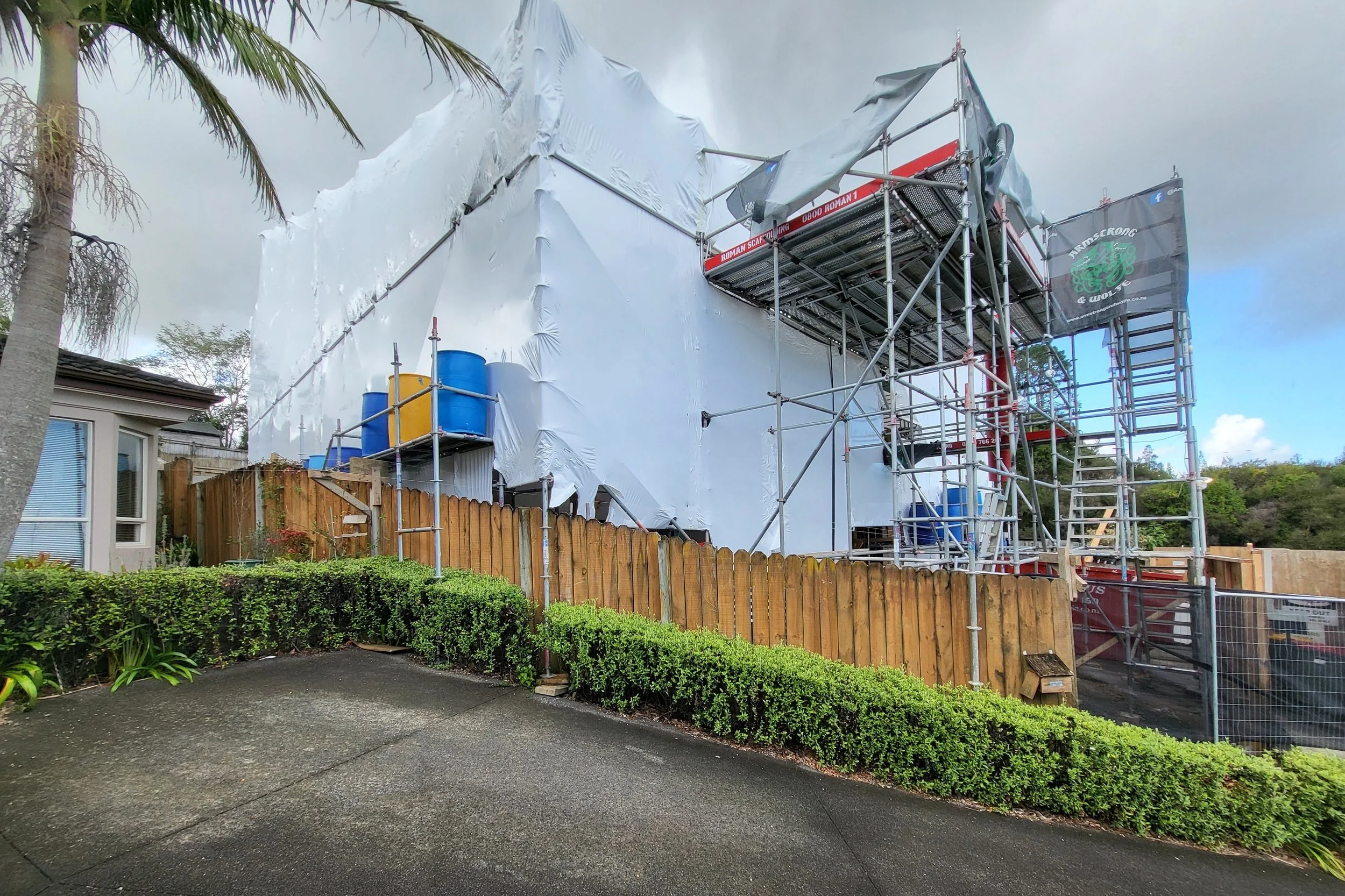 A house under construction or renovation with scaffolding around it. The house is wrapped in white plastic, and there are blue and yellow containers on the scaffolding. A wooden fence encloses the yard, and there are trees and bushes nearby with a cloudy sky overhead.